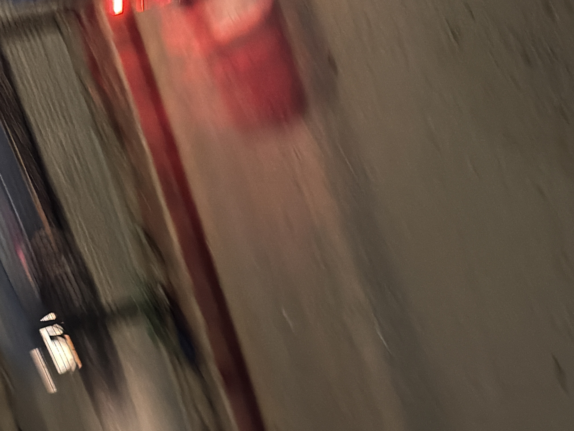 This is a motion-blurred photograph taken at night on a street in Bukuru, Nigeria. The image captures the movement of a vehicle, indicated by streaks of red light from its taillights and the blur of the surrounding environment. A portion of the vehicle's body, likely the rear, appears as a large, reddish-orange blurred mass on the right side of the frame. The street surface is visible as a textured, mottled grey-brown area, suggesting it is paved. Along the left and upper portion of the image, there are abstract streaks of light and dark, possibly representing streetlights or buildings. A thin red line, potentially a curb or painted road marking, runs horizontally across the middle of the image. The overall impression is one of rapid movement and low-light conditions.