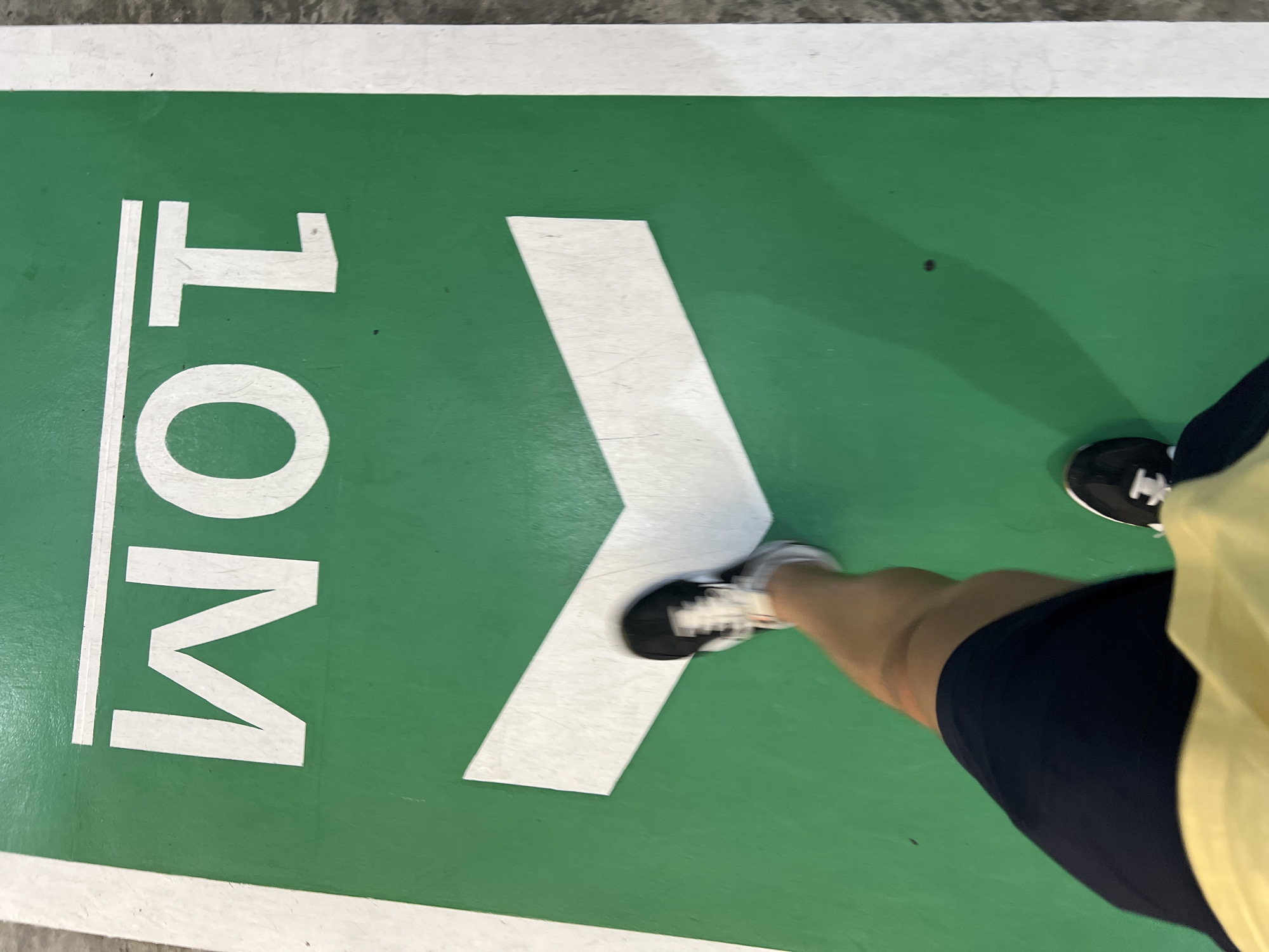 A high-angle, color photograph shows a green surface with a white directional marking of "10M" and an arrow. Part of a person is visible, wearing dark shorts, a yellow shirt, and black and white sneakers. The location is Unknown, Unknown.