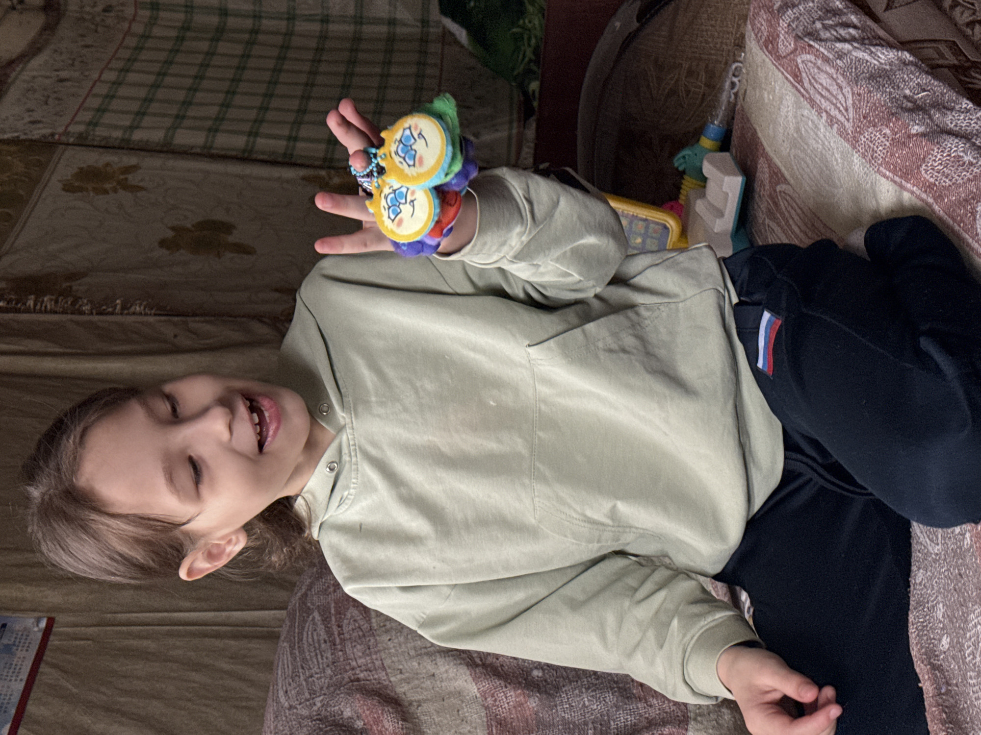 A young child with light brown hair is lying on a bed. The child is wearing a light green hoodie and dark blue pants. The child is holding a colorful toy in their right hand. The child is smiling and looking off to the left. The location is indoors, seemingly in a domestic environment. Based on a small flag on the child's pants, the possible location is Nizhnyaya Chermoda, Russia.