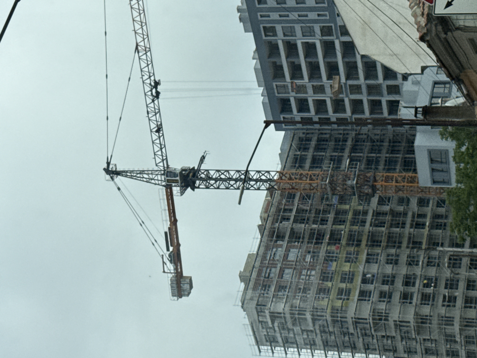 The image captures a bustling construction site in an urban setting, likely Varna, Bulgaria, under an overcast sky during daytime. The scene is dominated by two large tower cranes actively involved in building multiple high-rise structures.
In the foreground, a prominent tower crane features a long jib extending across the frame. Multiple small figures, identified as construction workers, are visible on its lattice structure; at least two appear to be climbing or working on the crane's boom, and another is positioned lower down near the operator's cabin.
Two multi-story buildings are under construction. One building, with a grey facade, has its concrete structure largely completed, featuring numerous open window and balcony frames. The second building is extensively covered in scaffolding, indicating an earlier stage of construction or facade development. On this heavily scaffolded building, at least two workers wearing yellow and white hard hats can be seen.
Several power lines or communication cables stretch horizontally across the sky between the crane and the buildings. On the far right edge of the frame, a partial view of an older building or structure is visible, along with a street sign displaying a black arrow pointing right. A small section of a green tree is also visible on the right. The overall impression is one of intense urban development and ongoing construction activity.