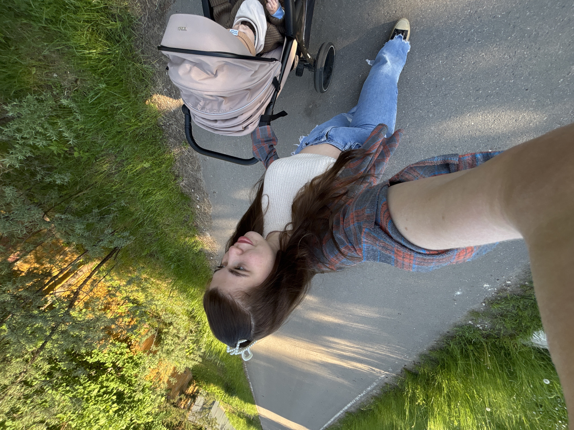 This is an outdoor selfie taken by a young woman pushing a baby stroller on a paved path, likely in Мещёры, Russia. The woman, with long, dark hair and fair skin, wears a white ribbed crop top, a blue and red plaid overshirt, and light blue ripped jeans. A decorative white, possibly pearl, hair clip is visible in her hair. She looks towards the camera with a subtle smile.
The beige-colored baby stroller, featuring text like "MIMU" and "LOVE WITH JOVOLA" (partially obscured), contains a baby whose feet and legs are visible, dressed in light brown clothing. The paved path is dark grey asphalt, flanked by vibrant green grass and various bushes and shrubs. Long shadows stretch across the path and vegetation, suggesting the photo was taken during late afternoon or early morning sunlight under clear weather conditions. The setting appears to be a park or a scenic walkway.
