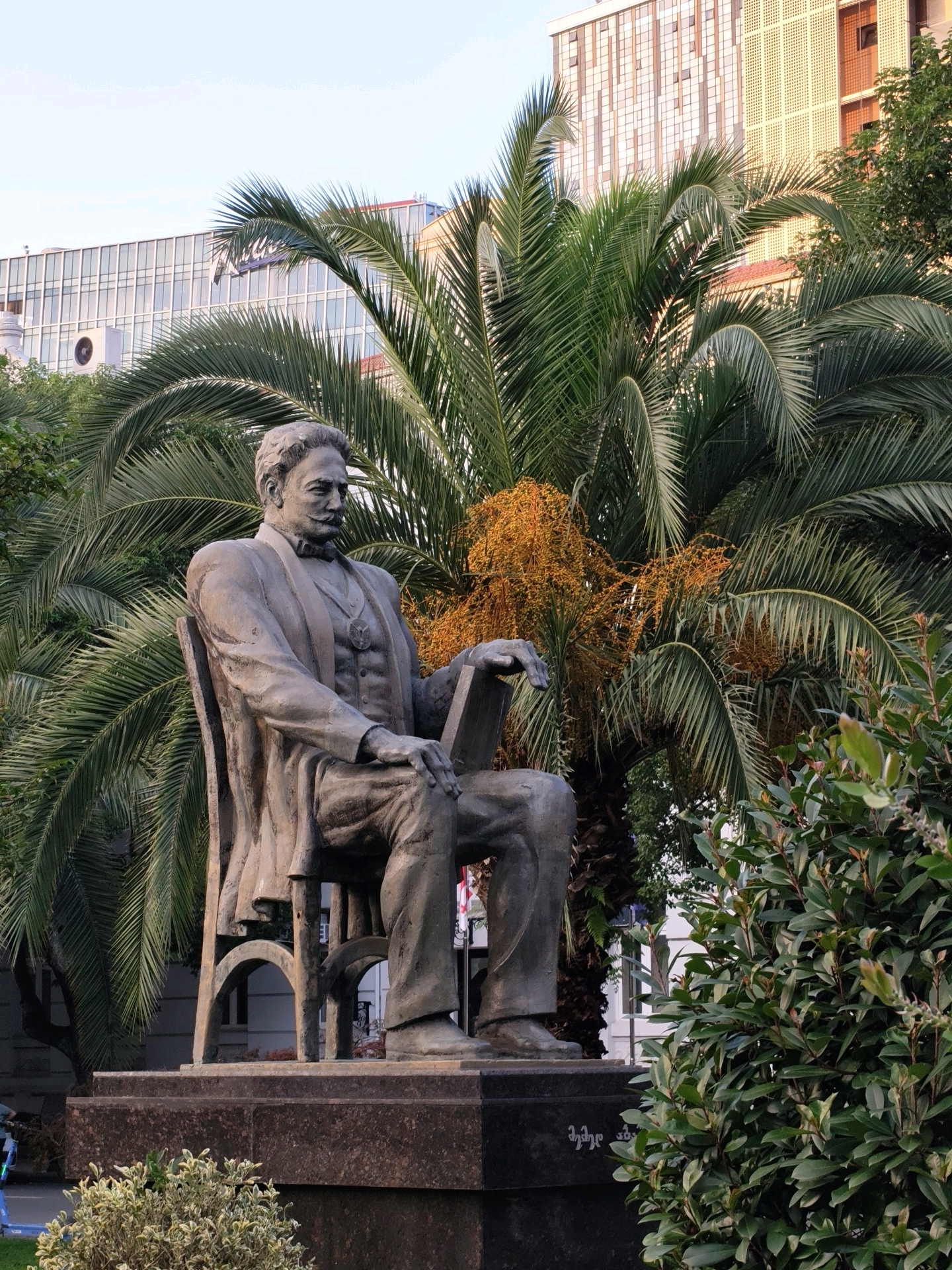 This is a ground-level, eye-level, vertical shot of a bronze statue of a seated man in Batumi, Georgia, likely in the Batumi Boulevard. The statue depicts a man with a mustache, wearing a suit, bow tie, and a medal on his chest, holding a book. He is seated on a chair with an open book resting on his lap. The statue is on a stone pedestal. Behind the statue, there are several large palm trees with dense foliage. The backdrop includes modern buildings with glass facades and a clear sky. In the foreground, there are green bushes and part of the pedestal. A small section of a Georgian flag is visible on the right side of the pedestal. The lighting suggests it is either early morning or late afternoon.