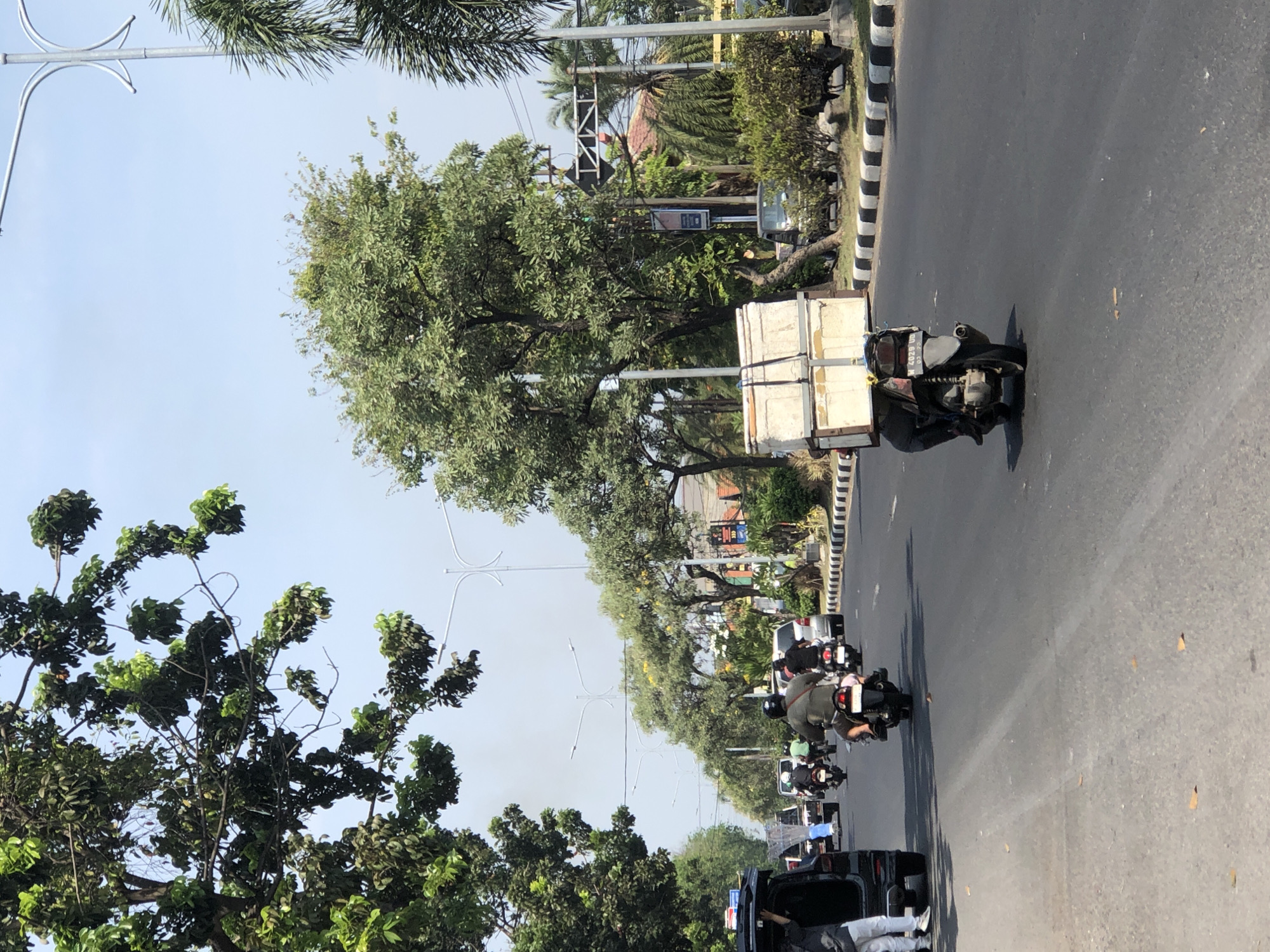 An outdoor, eye-level, medium shot shows a street scene in Larangan, Indonesia. The paved road has a black and white striped curb on the right side. Multiple trees line the street, with some branches extending over the road. Several vehicles are visible, including a black SUV on the left, a white van, and multiple motorcycles. A person is standing next to the black SUV. One motorcycle in the foreground is carrying a large, white, rectangular container secured with straps. Another motorcycle is behind it. In the background, there are more trees, buildings, and traffic lights. The sky is clear and blue.