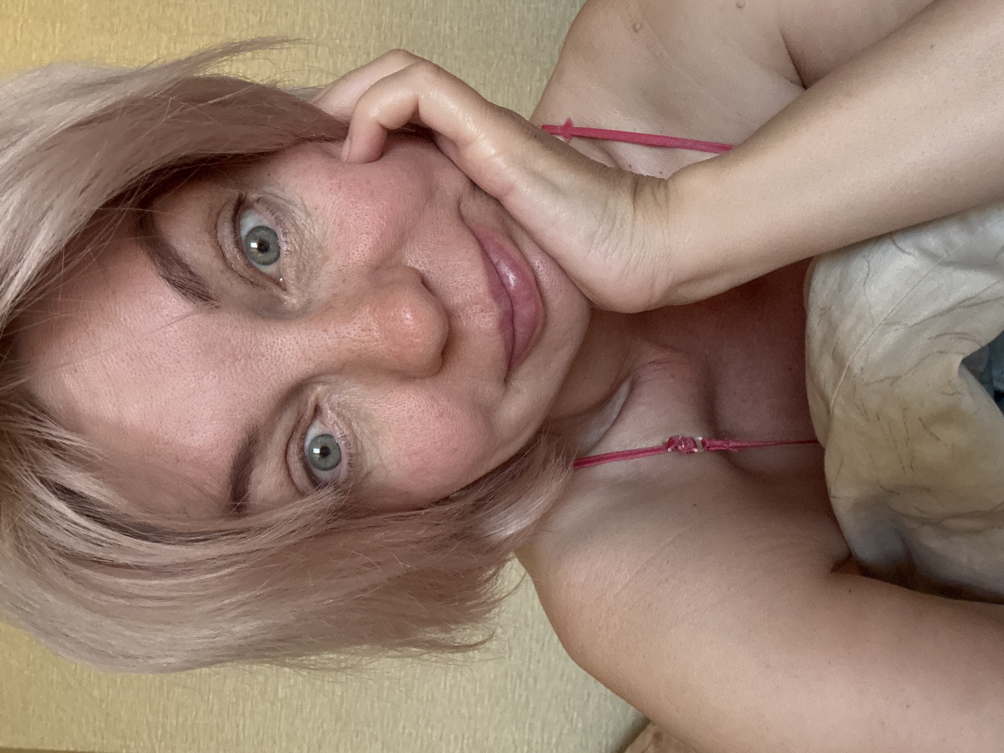 This close-up, sideways selfie captures an adult woman indoors, possibly in a bedroom or living room in Sochi, Russia. The woman has short, light pinkish-blonde hair that appears slightly disheveled. Her wide, striking light blue-grey eyes gaze directly into the camera, conveying a sense of directness and perhaps curiosity or playfulness. A subtle, soft smile plays on her lips.
Her left hand rests gently against her cheek and jawline. Her complexion is light, with visible natural skin texture and what appear to be freckles or subtle skin variations across her face. She is wearing a thin, pink strap of a top or bra, visible over her shoulder, and her bare shoulder and upper arm are prominent in the foreground, suggesting a relaxed or intimate setting. A light-colored pillow or blanket is visible to her right (bottom right of the image frame). The background features a plain, light yellow or beige wall, indicating a simple indoor environment. The lighting is bright and even, suggesting it is daytime. There is no visible text in the image.