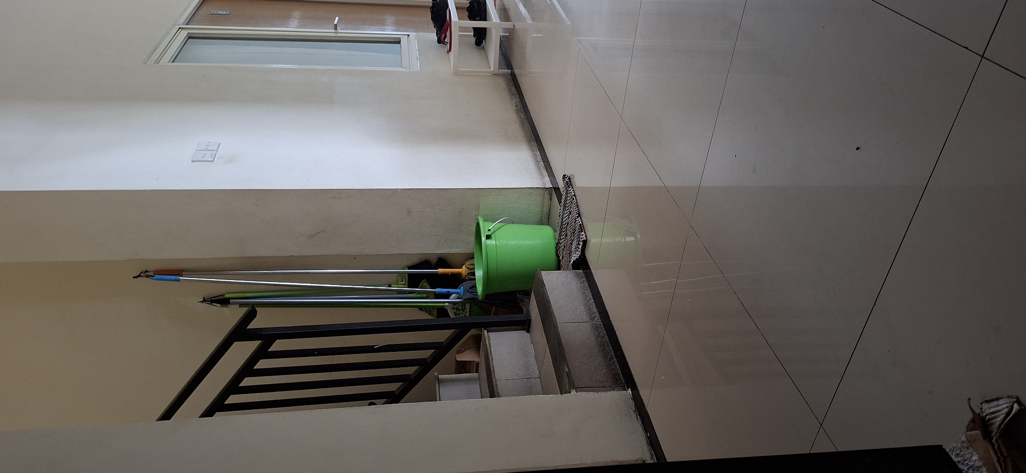 The image depicts an indoor scene, likely a hallway or entryway in a residential or commercial building in Surabaya, Indonesia. In the foreground, a polished, tiled floor reflects light. To the left, a staircase with black railings ascends, with concrete steps visible. Next to the stairs, a bright green plastic bucket stands on the floor, adjacent to a woven mat. Behind the bucket, several cleaning tools, including brooms and mops with colorful handles, are leaning against a cream-colored wall. Further to the right, a white shoe rack holds a pair of black shoes and a pair of red flip-flops. A doorway with a glass pane and a wooden door is visible in the background, along with a light switch on the wall. The lighting suggests it is daytime.