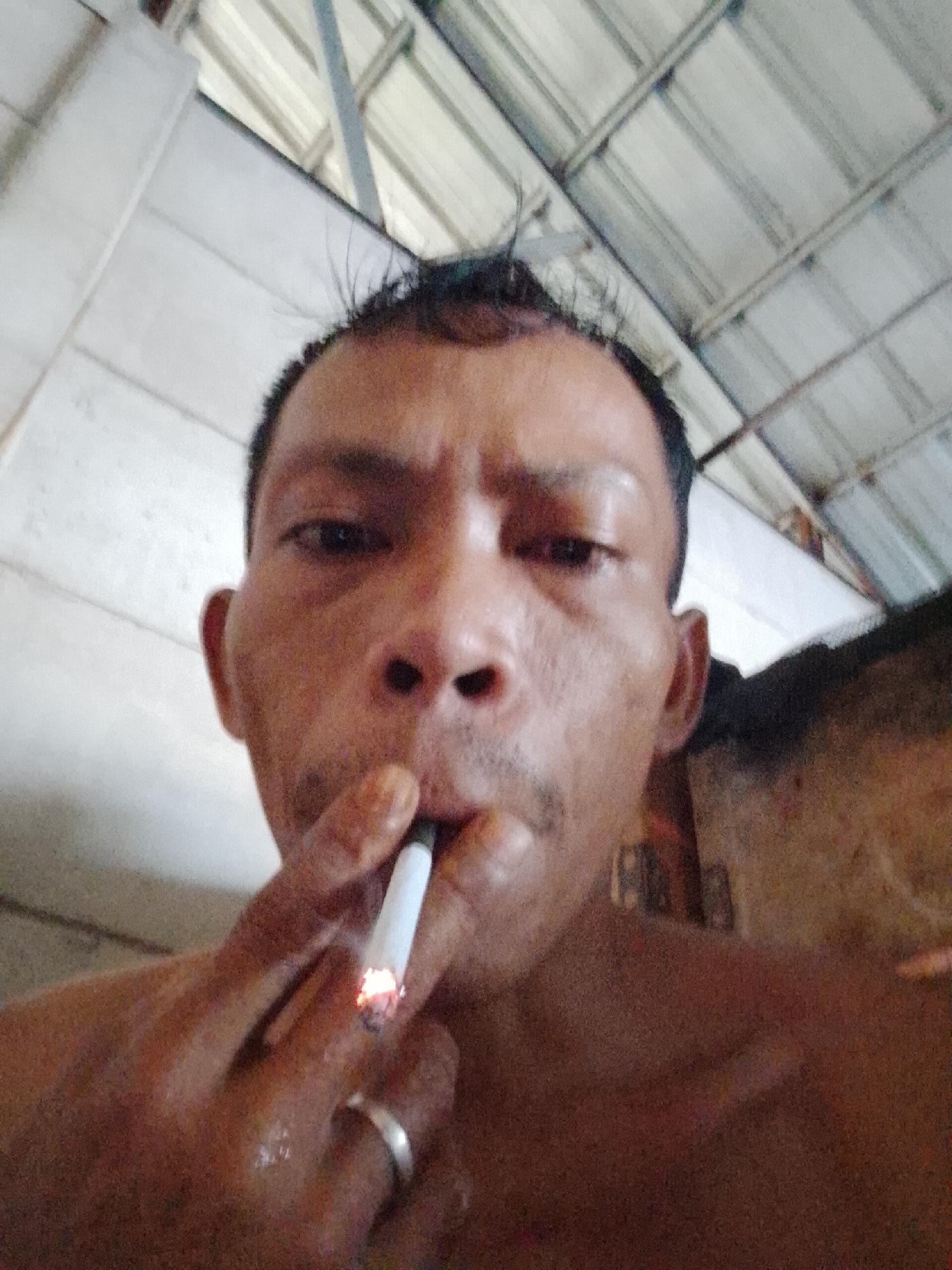 A close-up, low-angle shot depicts an adult male with dark hair and skin, holding a lit cigarette in his mouth. He is wearing a ring on his left ring finger. The background includes a metal corrugated roof and what appears to be part of a concrete structure. The location is Bukit Ketok, Indonesia.