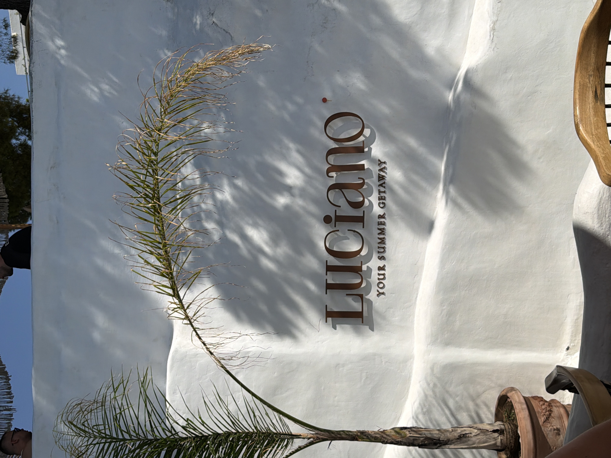 The image shows a white stucco wall with a sign that reads "LUCIANO YOUR SUMMER GETAWAY". The sign is made of dark, possibly bronze, lettering. To the left of the sign is a potted palm tree with a thin trunk and long, frondy leaves. The wall is textured and has shadows cast upon it from palm fronds and possibly other foliage. In the background, there are hints of a blue sky and some structures, possibly buildings with straw roofs. The lighting suggests it is daytime with bright sunlight. The style of the wall and the presence of the palm tree evoke a Mediterranean or coastal atmosphere. There are no visible people in the immediate foreground or interacting with the sign or the plant. The scene appears to be an exterior shot of a business or establishment. The provided crop images show that the location is in Himarë, Albania, and that there are two men visible in the background, looking over the wall. One man is wearing glasses and the other is wearing a black shirt. The overall impression is one of a vacation destination.