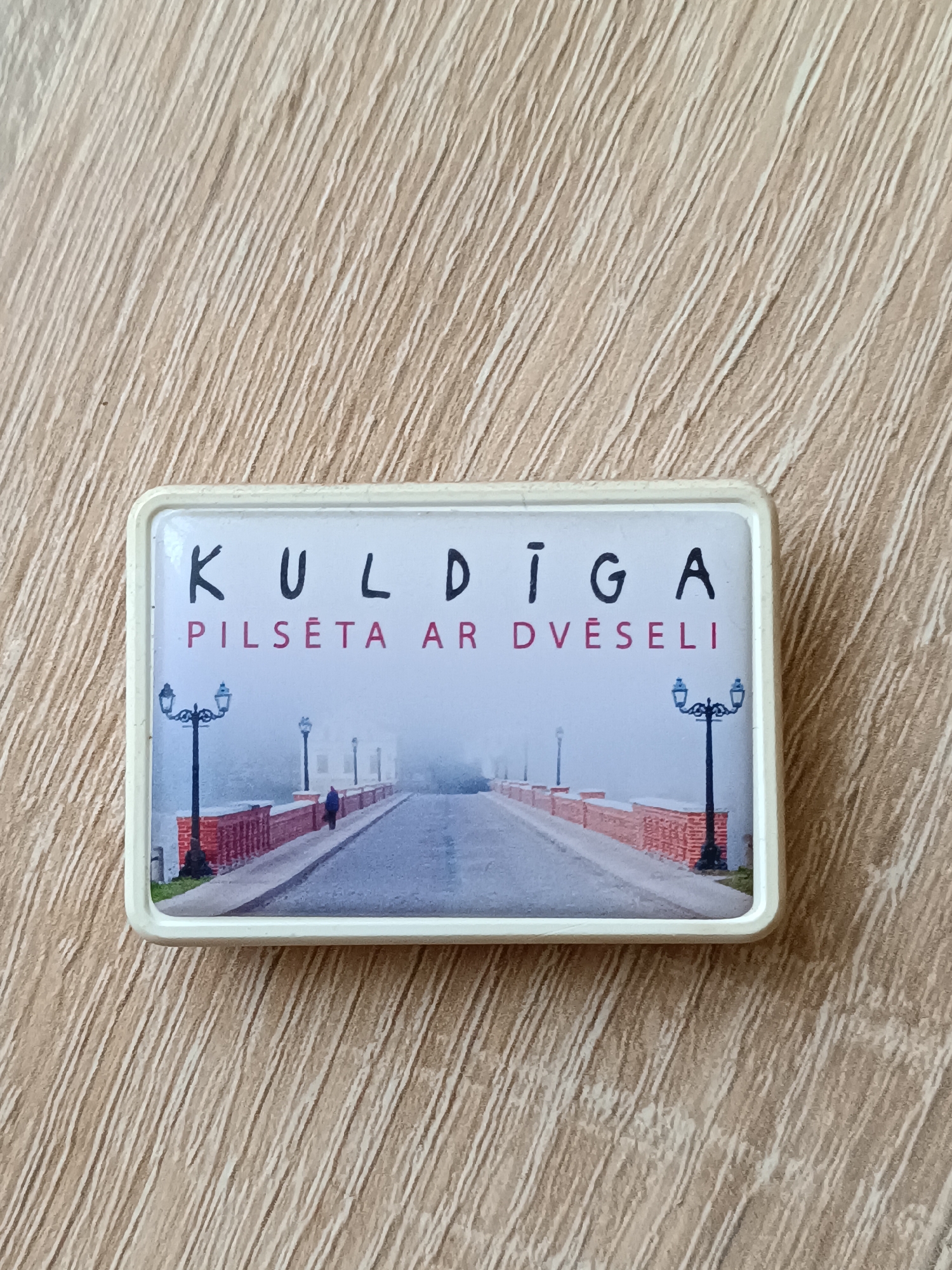 This is a rectangular magnet featuring a scenic photograph of a bridge. The magnet has a white border. The photograph depicts a street lined with red brick railings and old-fashioned lampposts. A lone figure, a person in a dark coat, is walking away from the viewer down the street. The scene is shrouded in a thick fog, suggesting a cool, misty atmosphere. The background is obscured by the fog, creating a somewhat mysterious or serene mood.
Above the image, the text "KULDĪGA" is printed in black capital letters. Below this, in smaller red capital letters, is the text "PILSETĀ AR DVĒSELI". The overall impression is a representation of Kuldīga, a town in Latvia, possibly highlighting its historic charm and tranquil ambiance, as suggested by the fog and the phrase "city with a soul". The time of day is ambiguous due to the fog, but the presence of lampposts might imply it's either early morning or late evening.