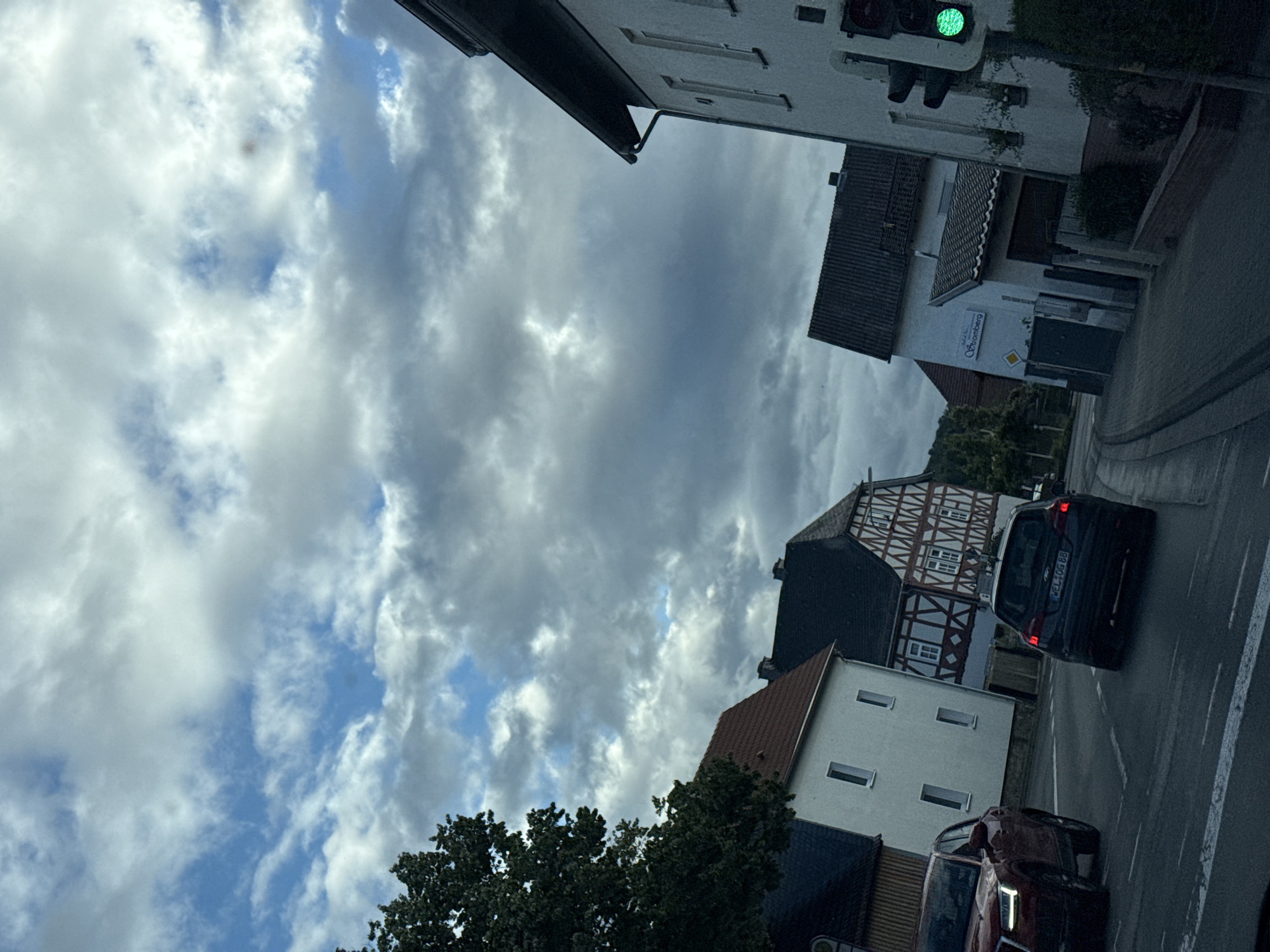 This is a photograph of a street scene in Volpertshausen, Germany. The image is oriented vertically, suggesting it was taken from inside a vehicle looking upwards and to the side.
The subjects include several buildings, a traffic light, and two cars. The buildings have traditional German architectural elements, with half-timbered facades visible on one structure. One building appears to be a business with signage that reads "Stromberg". The traffic light is green. The cars are a dark-colored hatchback and a red sedan. The red sedan's headlights are on, and the dark hatchback's taillights are illuminated.
The scene depicts a street with a paved road and a sidewalk. Trees are visible, with one large tree in the foreground. The sky is filled with dramatic clouds, suggesting cloudy or partly cloudy weather. The lighting indicates it is daytime, possibly late afternoon or early evening. The overall mood is neutral, capturing a typical street view.
There is visible text on the license plate of the dark hatchback, which appears to read "WEL-OG 88". The sign on the building reads "Stromberg". The traffic light has an illuminated green signal.