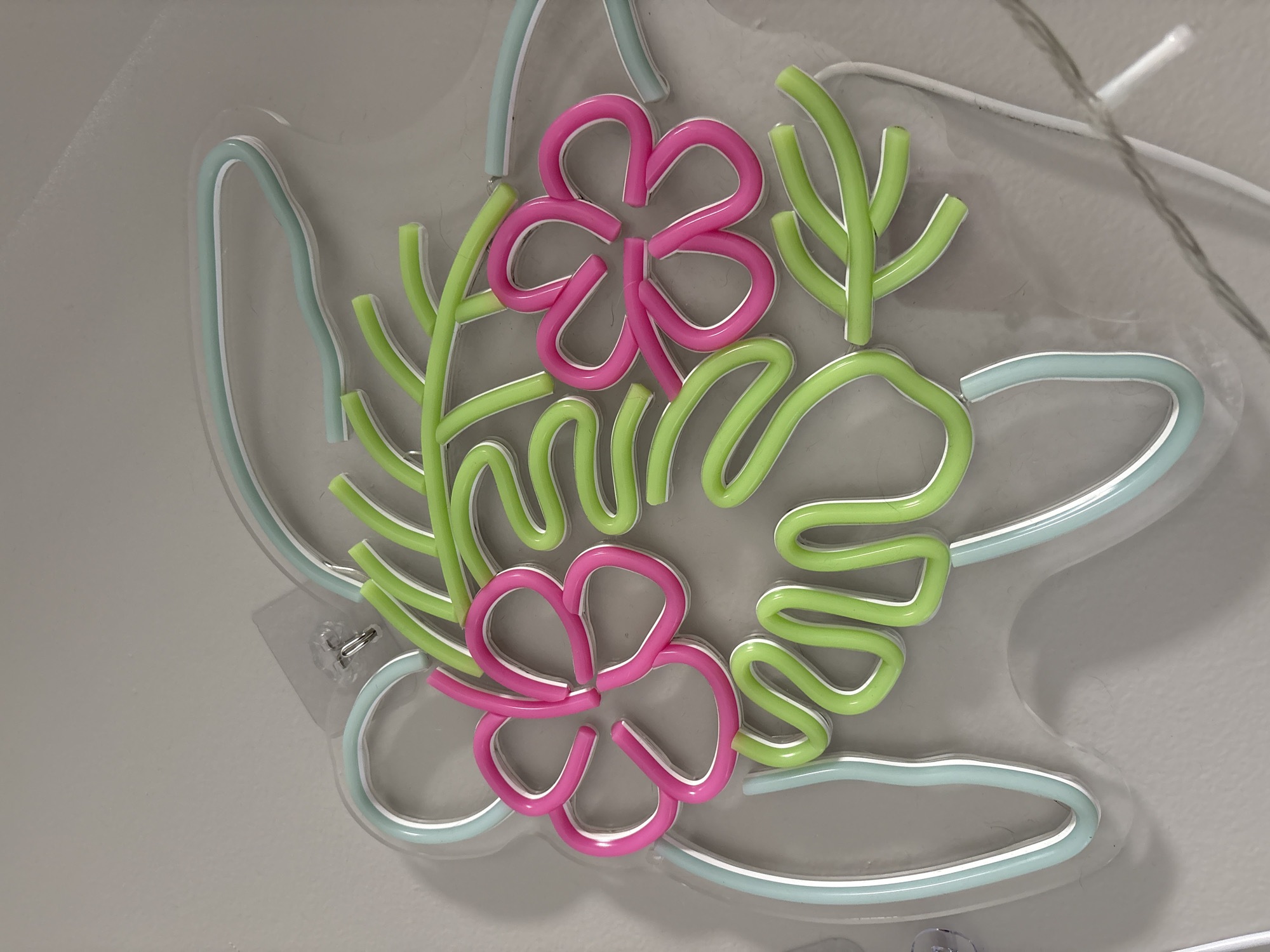 The image shows a neon sign mounted on a light gray wall. The sign features a stylized arrangement of flora. The sign's base is an amorphous shape outlined in pale blue neon tubing. Within this outline are two four-petal flowers formed by pink neon tubing, interspersed with greenery represented by green neon tubing shaped into stylized leaves and stems. The sign is attached to the wall with clear adhesive hooks. A thin, gray string is visible in the upper right corner. The location is unknown.
