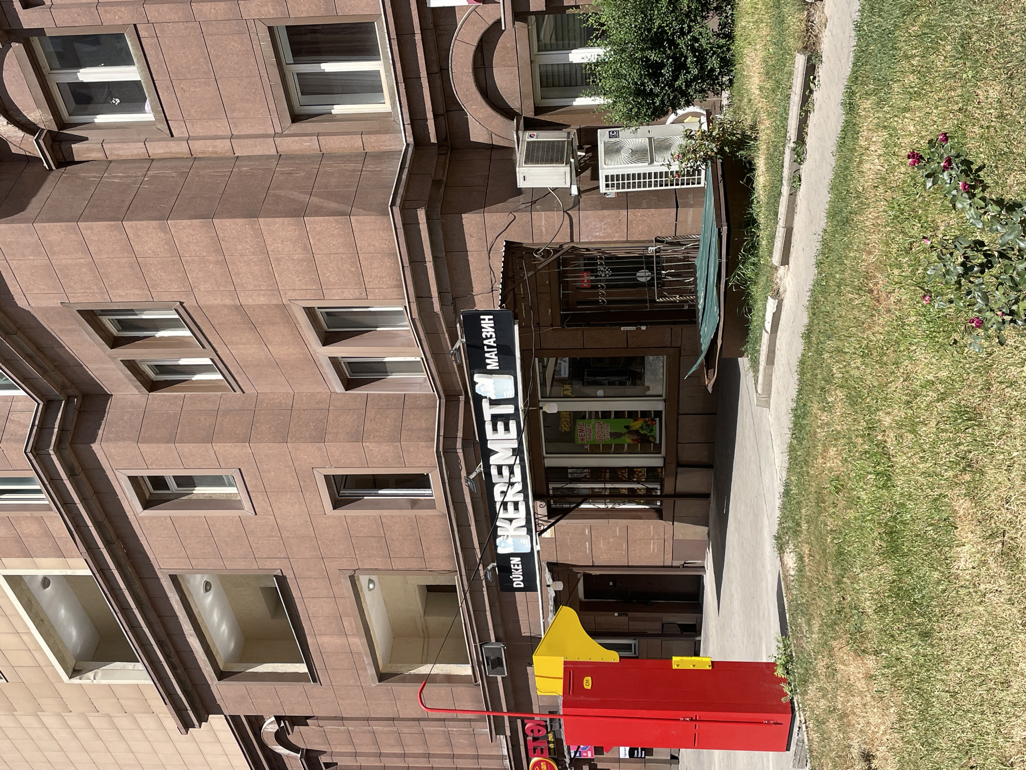 The image shows a storefront in Almaty, Kazakhstan. The storefront has a sign that reads "DÜKEN KEREMET МАГАЗИН" which translates to "Duken Keremet Store." There is a red metal cabinet and a yellow object on the left side of the image. The building is made of brown stone and has several windows. There are two air conditioning units mounted on the exterior wall of the building. In the foreground, there is a patch of grass with some small rose bushes. The sky is clear and the lighting suggests it is daytime.