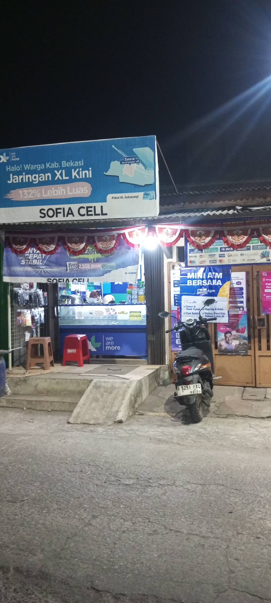 An outdoor, nighttime scene captures a street-level view of a mobile phone shop named "SOFIA CELL" in Setu, Indonesia. The shop features a blue storefront with advertisements displayed on its facade and above the entrance. A large blue banner announcing "Halo! Warga Kab. Bekasi Jaringan XL Kini 132% Lebih Luas" (Hello! Bekasi Regency Residents XL Network Now 132% Wider) is prominently displayed. Below this banner, the shop name "SOFIA CELL" is clearly visible.
Inside the shop, visible through the open entrance, there are shelves stocked with various items, likely mobile phone accessories and merchandise. Display cases and a counter area are also apparent. To the left of the entrance, a brown plastic stool stands on a concrete step. To the right of the stool, a red plastic stool with the word "SIMPATI" printed on it is placed near the shop entrance.
In front of the shop, a concrete ramp slopes down from the entrance to the street level. A black scooter is parked on the right side of the ramp, facing away from the viewer, with its license plate reading "B 5291 FXG". Further to the right, a portion of a yellow wooden door is visible.
The environment is dimly lit, with artificial lighting illuminating the shop and its surroundings. The ground is paved with asphalt, showing signs of wear and cracking. The overall impression is of a small, local business operating in a residential or commercial area at night.