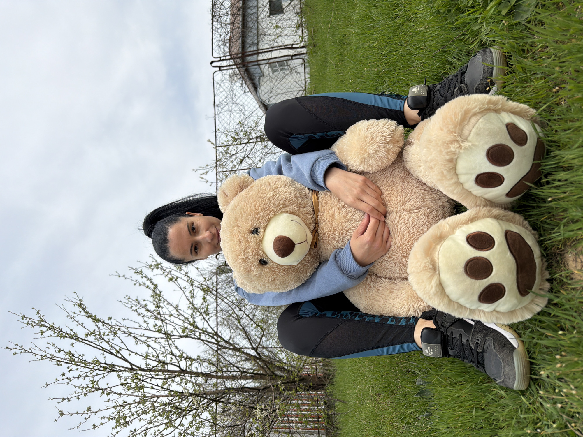 A woman with dark hair pulled into a bun sits cross-legged on a grassy area outdoors. She wears a light blue sweatshirt and patterned leggings, as well as black and gray tennis shoes. She holds a large, light brown teddy bear. A building with a wire fence sits in the background. There is a tree and cloudy skies in the background. The location is Pleven, Bulgaria.