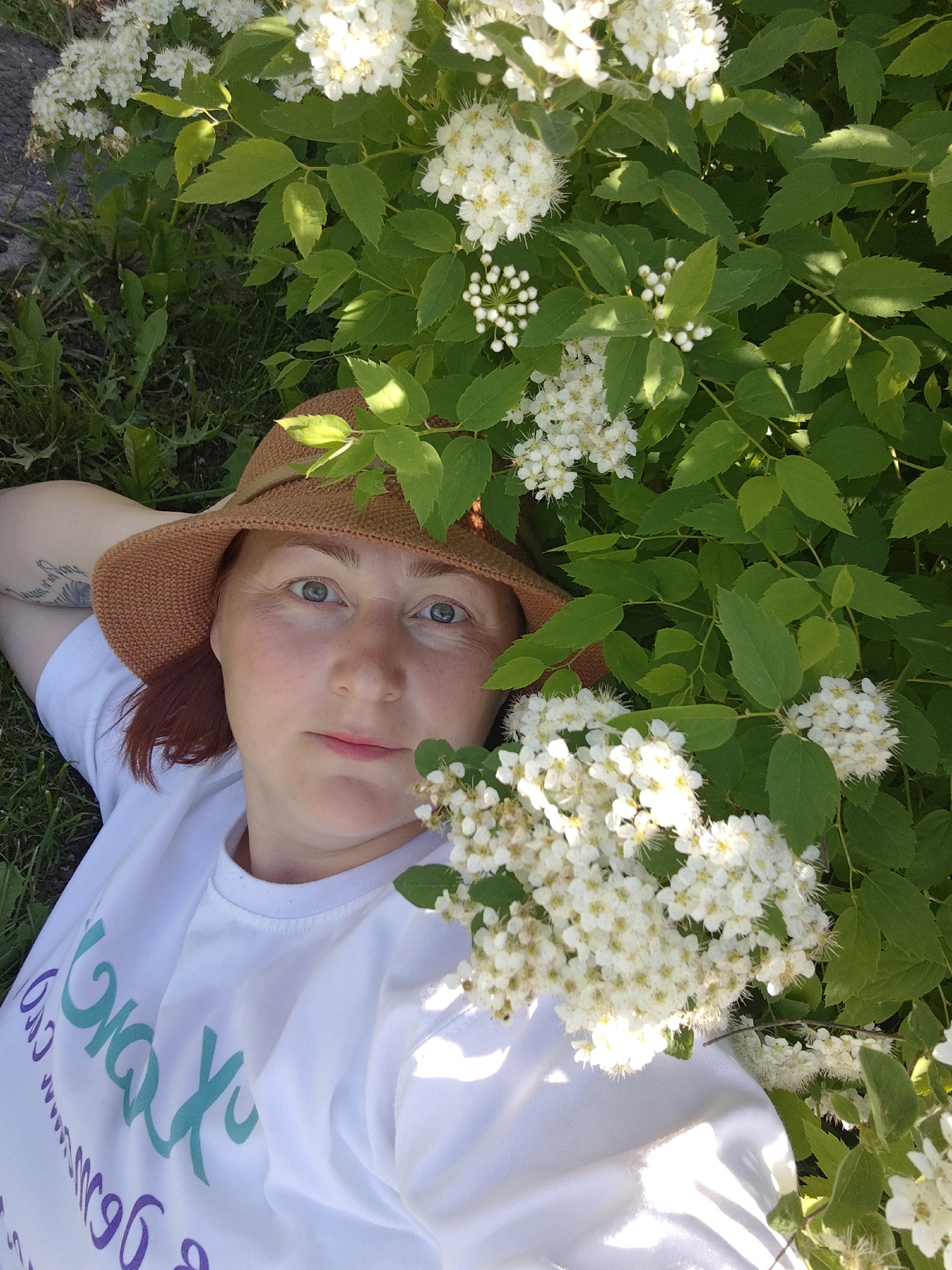 A woman is lying in a grassy area with flowering plants. She is wearing a light brown woven hat and a white t-shirt with writing in teal and purple. Her arm is raised behind her head, and a tattoo is visible. The woman is looking directly at the camera. The flowering plants feature green leaves and clusters of small white flowers. Some grass is visible to the left of the woman. The location is Фоки, Russia.