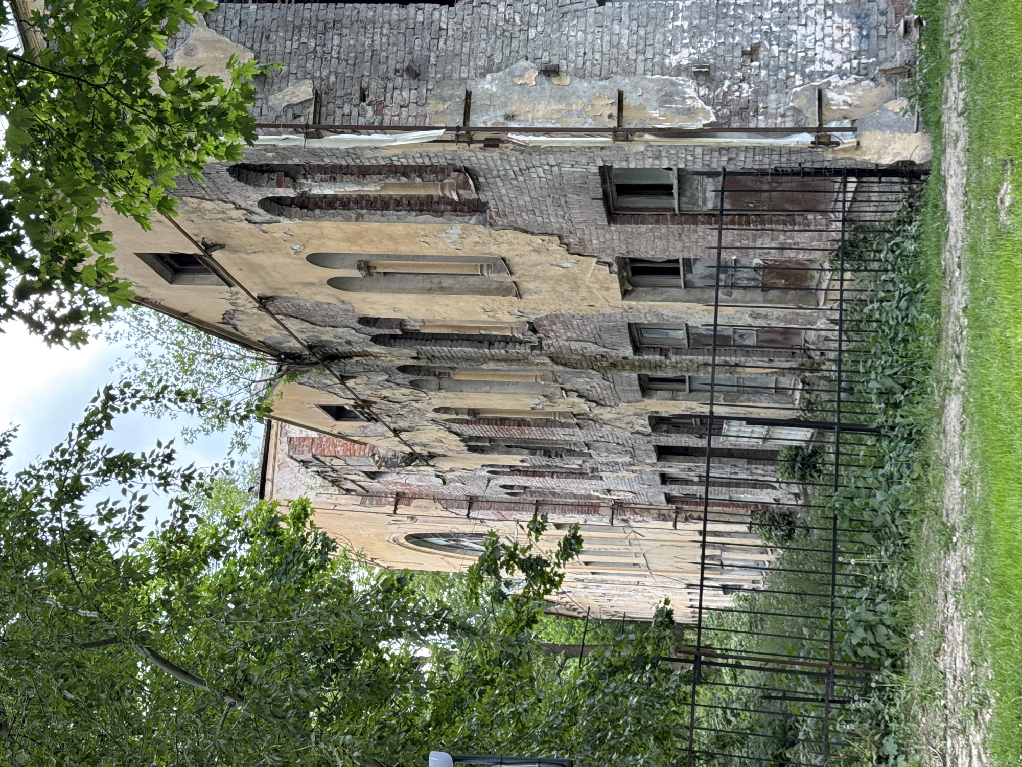 This image depicts a severely dilapidated, multi-story building, likely located in Saint Petersburg, Russia. The building's facade is extensively decayed, with large sections of its original plaster or stucco peeling and crumbling away, revealing the underlying red-brown brickwork. This exposed brick contrasts with the remaining patches of lighter, faded yellow or beige facade material.
Numerous windows are visible across the building; some are dark, empty openings, others appear to be boarded up or broken, while one higher window retains a dark frame. Several arched recesses are partially discernible on the facade, suggesting original architectural details. Horizontal metal bars or supports are affixed to the upper portions of the building, possibly for structural reinforcement or remnants of past construction work.
In the foreground and left side of the image, lush green foliage from trees frames the scene, indicating a spring or summer season. A black metal fence runs along the front of the building, with wild green plants and weeds growing abundantly at its base, further emphasizing a sense of neglect. To the right of the fence, the ground consists of a strip of vibrant green grass adjacent to a lighter, dirt path or area. The sky, visible through the tree canopy, appears bright and overcast, suggesting daytime.
No people or activities are visible, contributing to a static and quiet atmosphere. The overall impression is one of an aged historical structure in significant disrepair, set against a backdrop of natural overgrowth. No visible text is present in the image.