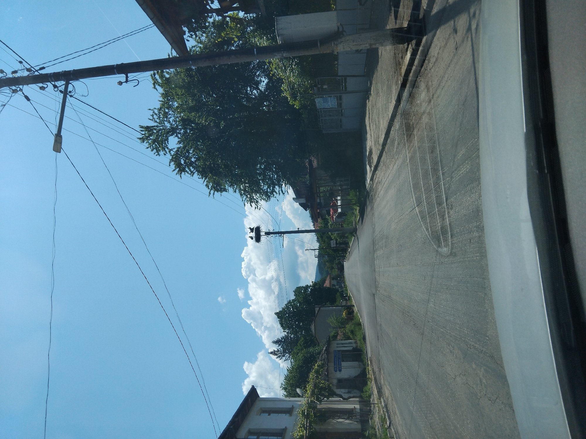 The image shows a view from inside a vehicle looking out onto a street in Velchevo, Bulgaria. The road is paved and appears to be a residential street. Utility poles are visible along the side of the road with power lines running overhead. A stork nest is located atop one of the poles. Houses and trees line the street. The sky is blue with scattered white clouds. The focus is on the street and the immediate surroundings.