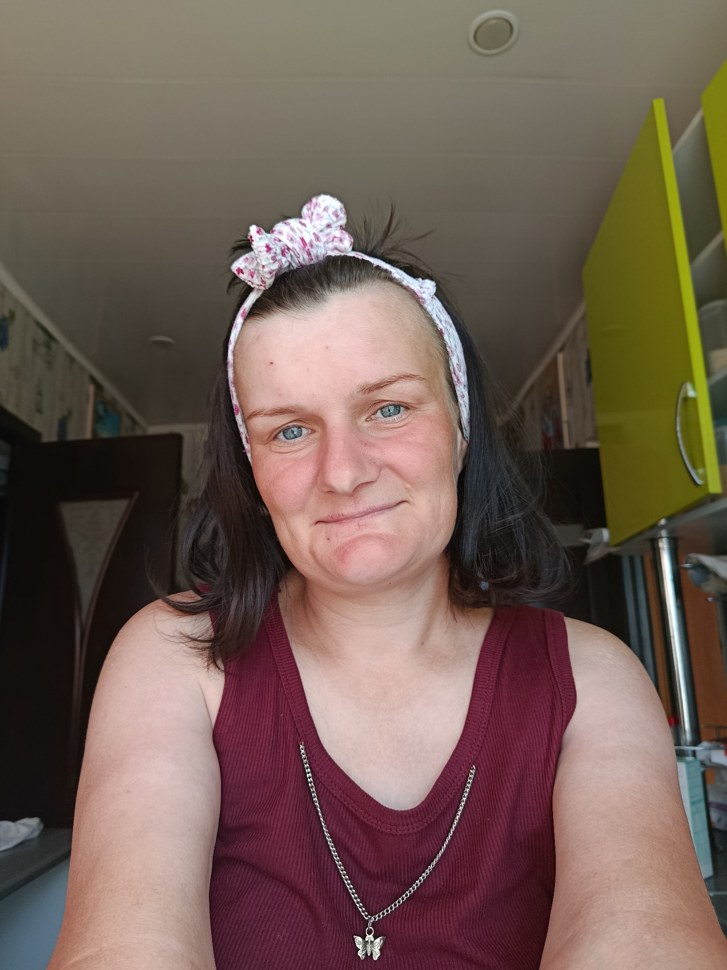 A middle-aged woman with short, dark hair is centered in the frame, wearing a maroon tank top and a floral headband. She has blue eyes and a slight smile. A silver necklace with a butterfly pendant hangs around her neck. The background suggests an indoor setting, possibly a kitchen, with yellow cabinets visible on the right. A door frame is seen on the left, revealing a glimpse of patterned wallpaper. The lighting appears natural, casting shadows that add depth to the scene. The location appears to be Фоки, Russia.