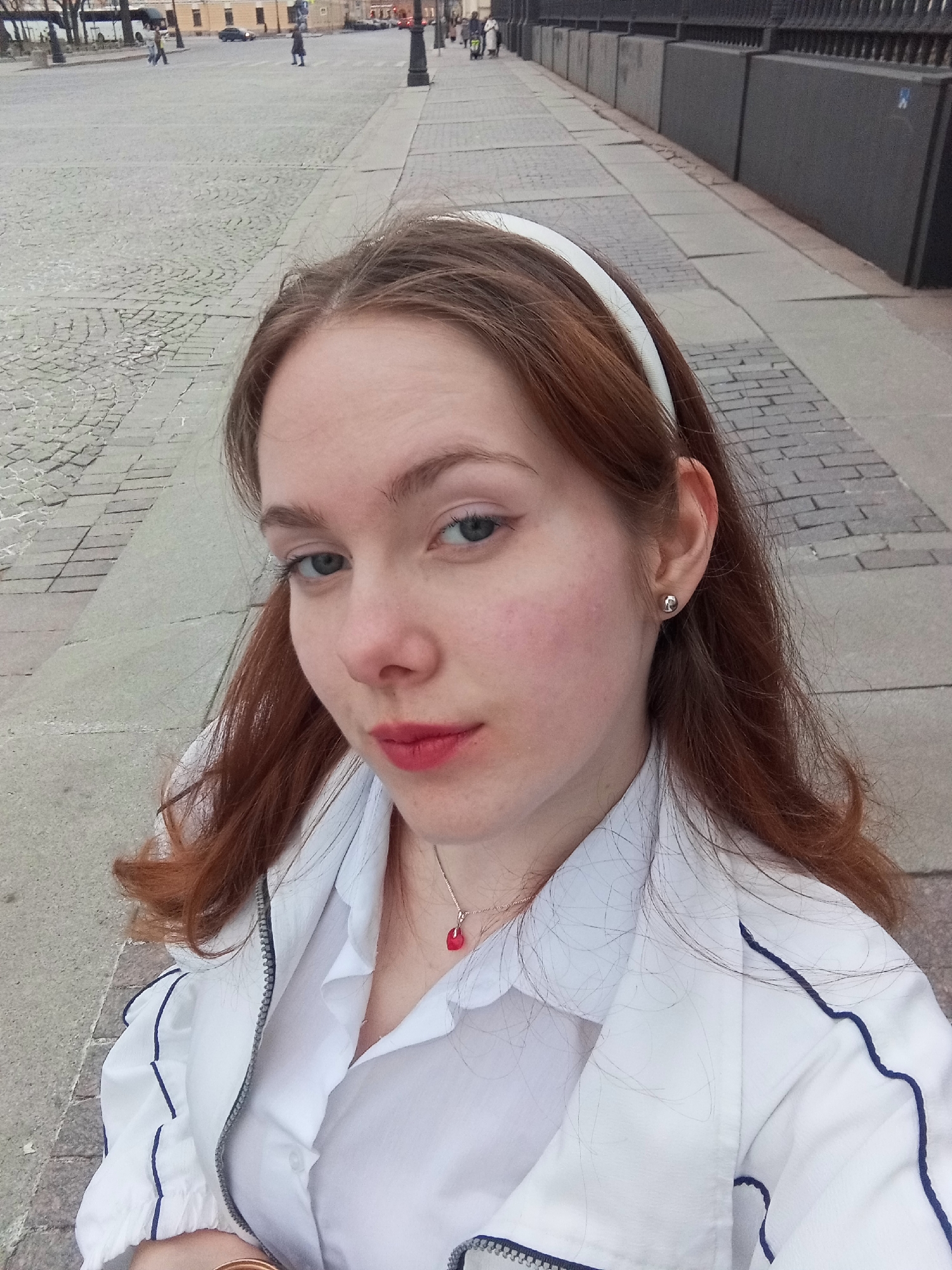 A woman poses for a selfie. She has fair skin, reddish-brown hair held back by a white headband, and blue eyes. She wears a silver necklace with a red pendant and silver stud earrings. Her lips are painted with a red lipstick. She is wearing a white jacket with navy blue trim over a white collared shirt.
She is standing on a paved area with irregular stone blocks. In the background, there is a street with a few people walking in the distance and buildings on either side. To the right, a dark grey fence runs along a sidewalk. Based on the architecture and street paving, the location is likely in Saint Petersburg, Russia.