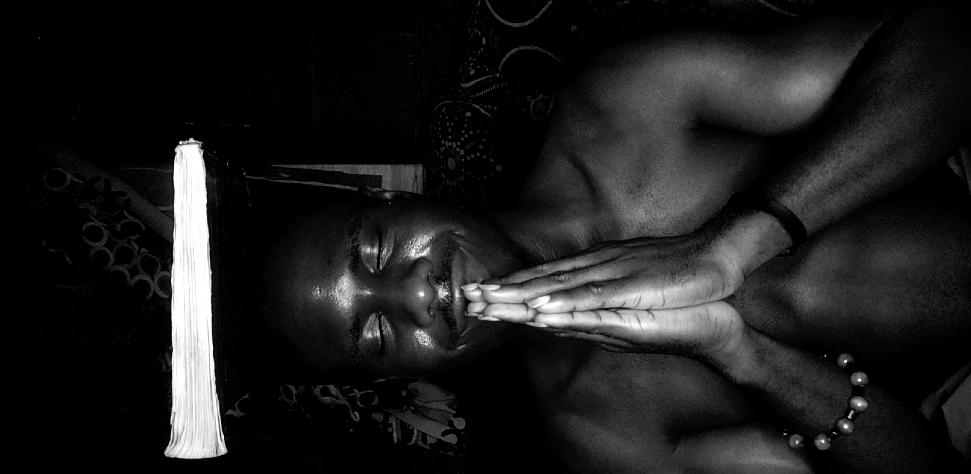 The image shows a shirtless man lying on his back, hands clasped in front of his face. He wears a bracelet and a black wristband on his left arm. A tall, thin white object stands to his left. The location is Narayi, Nigeria.