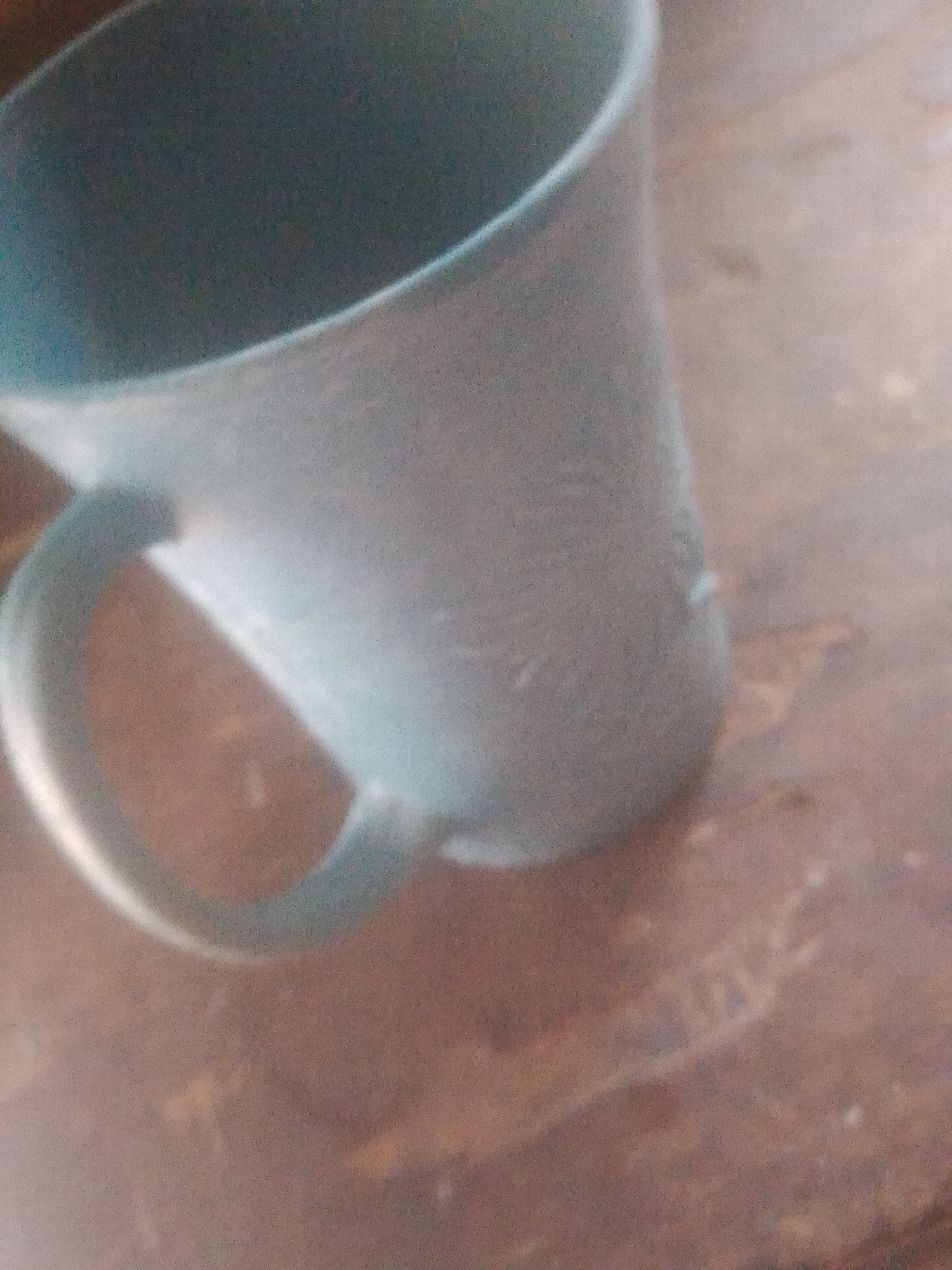 A light blue, slightly opaque mug with a handle is placed on a wooden surface in Jauro Yinu, Nigeria. The mug appears to be made of plastic and has a smooth texture. The wooden surface is a medium brown color with visible grain patterns. The lighting is dim, casting soft shadows. There are no people or any other objects visible in the frame. The image is focused on the mug and its immediate surroundings, with a shallow depth of field blurring the background.