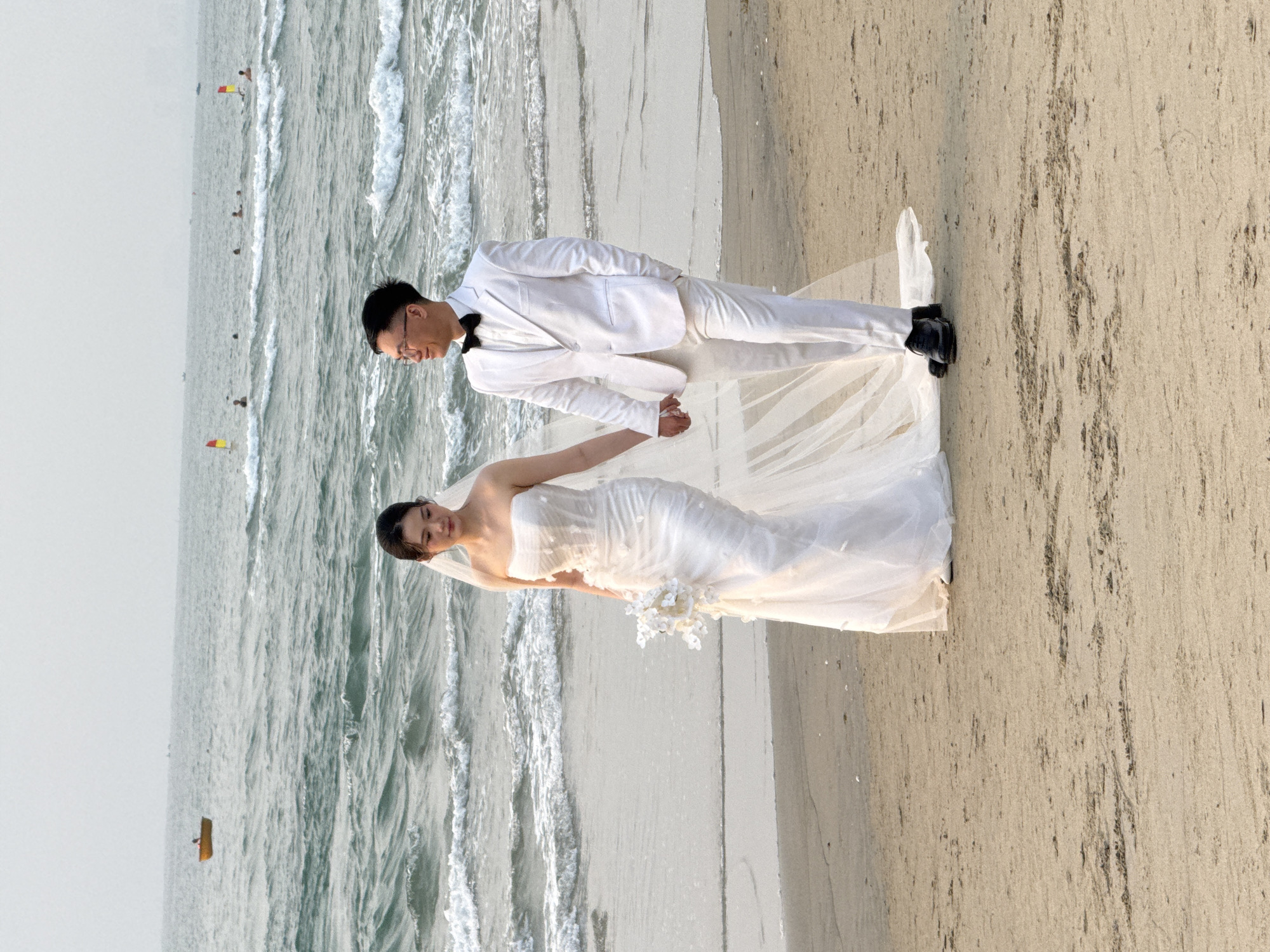 A wedding photo shows a bride and groom standing on a beach, holding hands. The bride wears a white, strapless gown and veil, holding a bouquet of white flowers. The groom wears a white suit, a bow tie, and black shoes. The backdrop includes the ocean with waves and a sandy beach. The location is Đà Nẵng, Vietnam, as indicated by the beach setting and possible visual cues of the region.