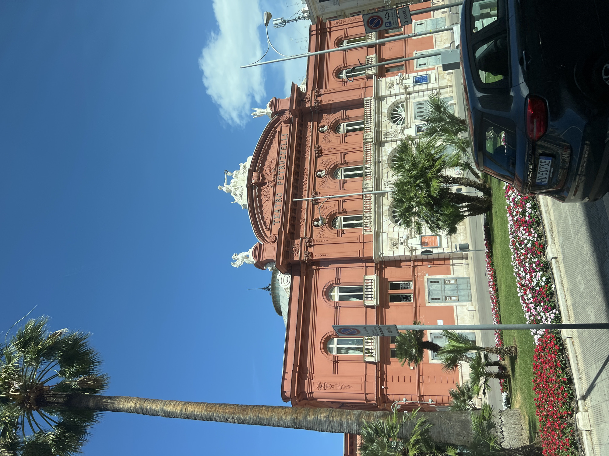 This image shows the Teatro Petruzzelli in Bari, Italy, on a clear day. A tall palm tree dominates the left side of the frame, with its fronds reaching towards the bright blue sky. In the center, the large, salmon-pink facade of the opera house is visible, featuring ornate architectural details, arched windows, and statues. The name "TEATRO PETRUZZELLI" is inscribed above the main entrance. In front of the building, a well-maintained garden bed is filled with red, pink, and white flowers. Several smaller palm trees are scattered throughout the garden. A dark-colored car is parked in the foreground, its rear visible to the viewer. The street in front of the garden has a paved surface. A street lamp and traffic signs are also present. The scene appears to be a public space, likely a street or plaza adjacent to the theater.