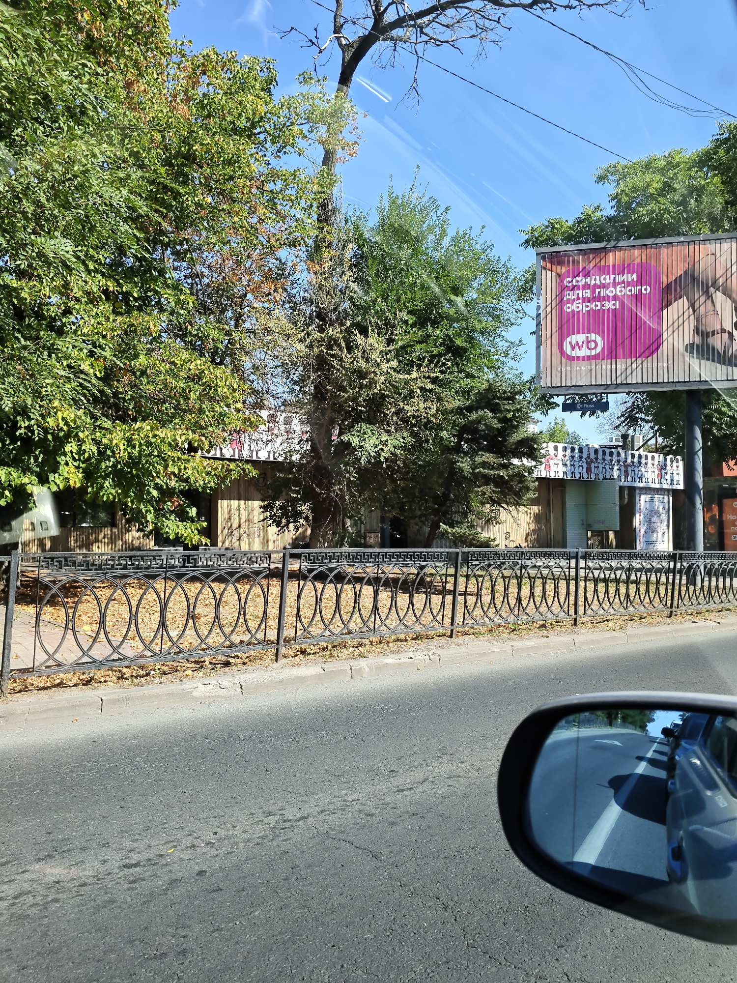The image captures a street scene in Rostov-on-Don, Russia, on a sunny day. The foreground shows a paved road with a car's side mirror reflecting other vehicles. Behind the road, a black wrought-iron fence with an intricate circular and geometric pattern separates it from a sidewalk covered with fallen leaves. Beyond the fence, there is a low building with a wooden facade and a row of white figures, resembling toy soldiers, displayed on its roof. Large trees with green and some autumn-colored foliage line the area behind the building. Further in the background, a large billboard with a pink background and text in Cyrillic, advertising "sandals for any look," is visible. The sky is clear blue with faint white contrails from an aircraft.