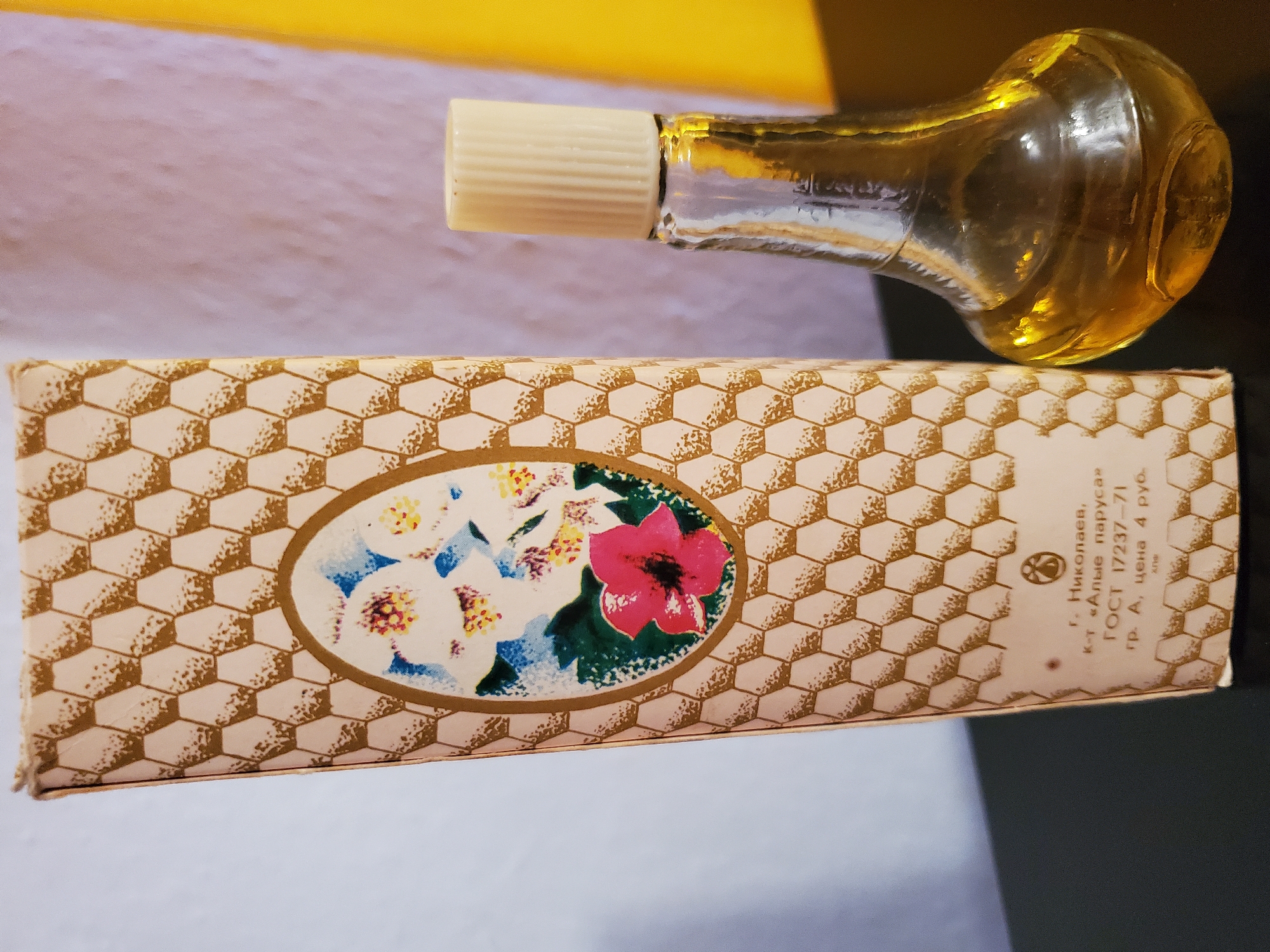 The image shows a small, vintage perfume bottle with a yellowish liquid inside and a ribbed, cream-colored cap. It stands next to a decorative cardboard box that features a honeycomb pattern and an oval inset with a floral illustration, which includes pink, blue, and yellow flowers. The box also has text printed on it in Cyrillic script. The scene is set against a background that appears to be a wall, with a bright yellow section on the right and a lighter, textured section to the left. There is also a dark surface in the foreground. The image is a still life composition, with no people or discernible actions. The bottle and box are positioned on a dark surface. The city of Konstantinovka, Moldova is not depicted or implied by the visual elements in the image.