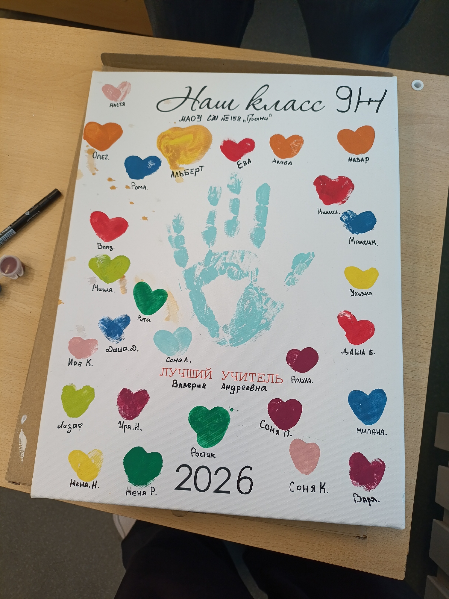The image is a photograph of a white canvas propped up on a wooden surface. On the canvas is a painted artwork. The artwork includes a title in Cyrillic script: "Haw knacc 9H" followed by "MAOY CAI No158 'Tpamu'". Below this title is a series of heart-shaped paint marks, each accompanied by a name written in Cyrillic. These names include: "HASTYA", "ΟΛΕΣ", "POMA", "ABBEPT", "EBA", "ANICA", "HA3AP", "Bnag", "Muwa", "MUMa", "Puta", "Hukuta", "MakcUM", "Иpa K.", "Aawa.A", "Covn.A.", "Ynbana", "AYuuu YMUTENb", "Aawa B", "Auuna", "Лn3ap?", "MUNAHA", "Hepa H.", "CoHA П.", "Upa H.", "POCTNK", "CoHA K.", "Bapa.", "Her R." and "2026." In the center of the canvas is a blue handprint made with paint. Beside the left edge of the canvas is a black marker pen, paint, and a jar.