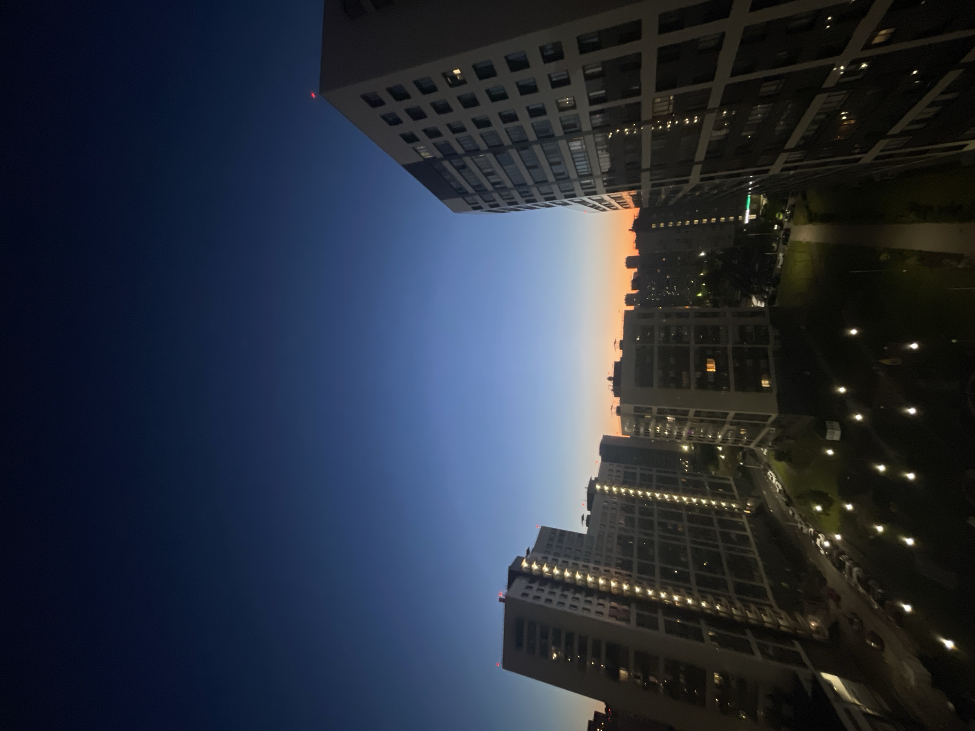 The image depicts a nighttime cityscape in Sinyavino, Russia, likely taken from an elevated perspective such as a balcony or window. The sky is a deep, clear blue, transitioning to hues of orange and yellow near the horizon, indicating either dawn or dusk. Multiple modern apartment buildings dominate the scene, their windows illuminated with warm, artificial light, suggesting occupancy. The architecture is characterized by repetitive rectangular windows and balconies, a common feature of contemporary residential construction. The street level is visible between the buildings, with parked cars lining a road and pathways crisscrossing grassy areas. Numerous small, illuminated outdoor lights are scattered across the ground, casting a soft glow. A red light is visible atop one of the taller buildings, likely a navigational light for aircraft. The overall impression is one of an active, urban residential area at twilight.