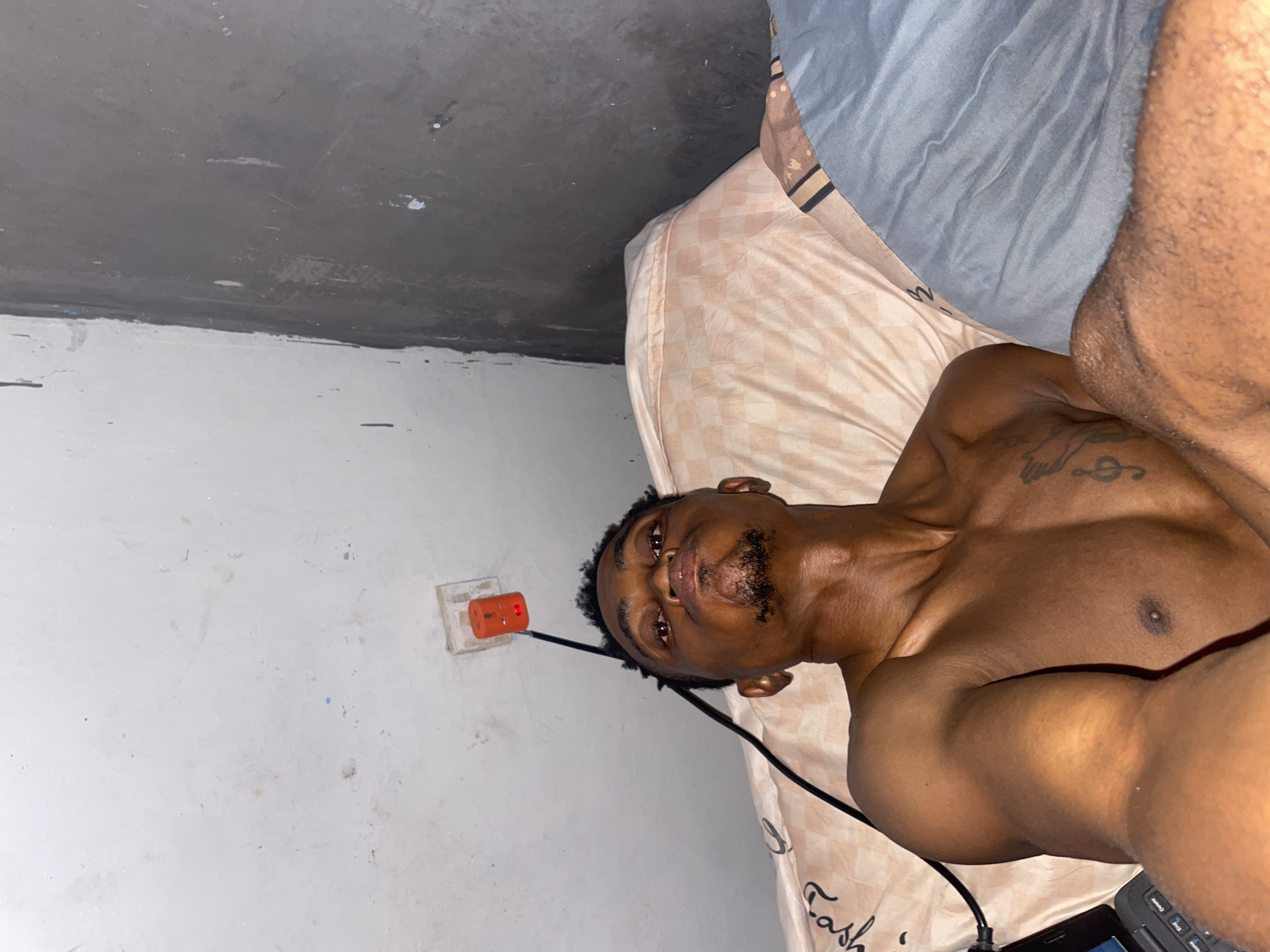 The image shows a young Black male lying on a bed. He is shirtless and appears to be in a bedroom. The room has a white ceiling with a light fixture and an orange electrical outlet. The walls are partly white and partly a dark, textured material. He is lying on a peach-colored pillow and what appears to be a blue duvet. A laptop is visible on the left side of the bed. The man is looking up towards the camera with a pouting expression. He has a short beard and a tattoo of musical notes on his chest. The scene is dimly lit, suggesting it may be nighttime or the room is not well-lit. The presence of a bed, laptop, and electrical outlet suggests a domestic or personal space. The location of Adankolo, Nigeria, implies a specific geographic and cultural context for the scene, though no direct visual cues in the image confirm this location.