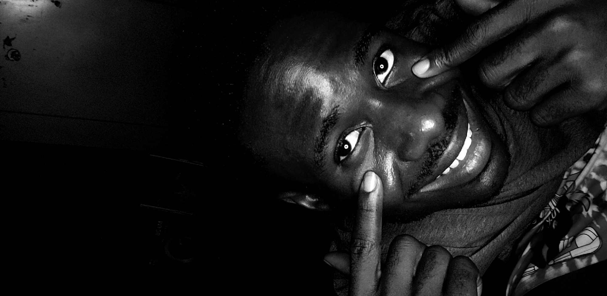 A striking black and white portrait captures a young adult male looking directly at the camera with a playful and exaggerated expression. He is smiling broadly, revealing bright white teeth, and uses both index fingers to pull down the skin under his eyes, making them appear wide and prominent. His eyes are bright and focused.
The subject's face is intensely lit by a frontal light source, creating strong highlights on his forehead, nose, cheeks, and lips, and emphasizing the texture of his skin. The background is overwhelmingly dark, suggesting either a low-light environment or a focused lighting setup that isolates the subject.
On the left side of the dark background, a faint object is visible, displaying discernible white text that reads "W 20 CM" and includes a partial circular dial or knob. To the right of the subject, a portion of a patterned garment, possibly a shirt or cloth with an abstract or geometric design, is visible, adding a subtle touch of detail to the edge of the frame. The overall mood is one of lightheartedness and direct engagement, captured in a high-contrast monochromatic style.