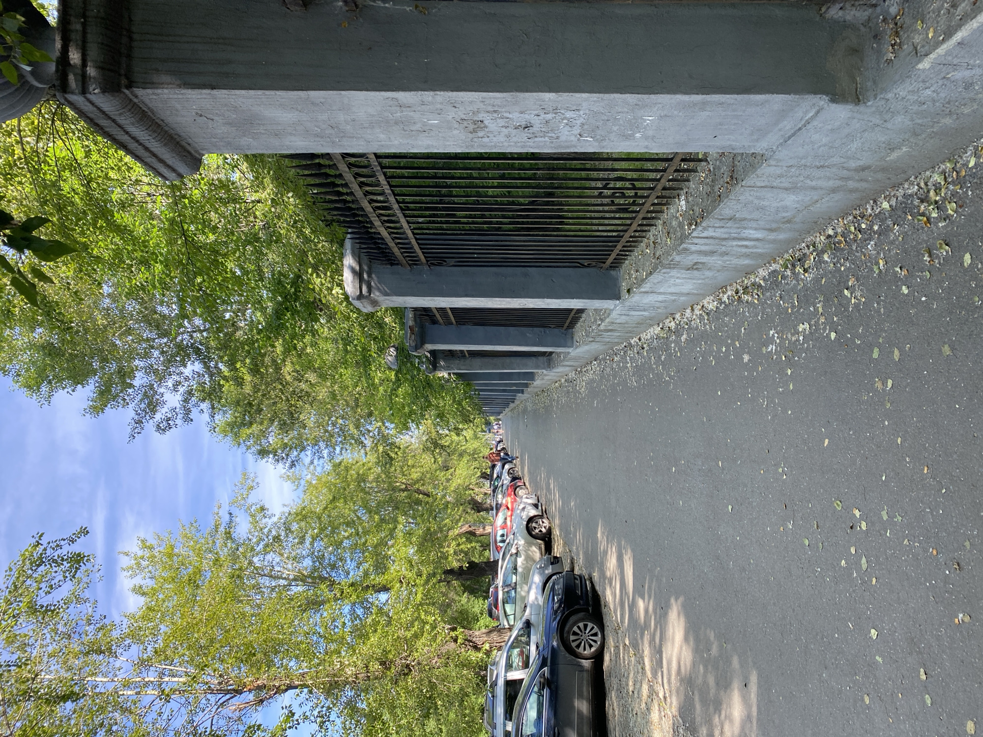 The image depicts a street scene in Yekaterinburg, Russia, likely during daylight hours given the bright illumination and shadows. The perspective is from ground level, looking down a paved walkway adjacent to a black metal fence supported by concrete pillars. The fence encloses an area with greenery, likely trees and bushes. To the left of the walkway, a row of parked cars is visible, suggesting a street or parking area. Trees with lush green foliage line the street on the left, casting dappled shadows on the walkway. The sky is visible in the upper portion of the image, appearing partly cloudy with a blue hue. A single person, appearing to be a man, is walking away from the camera on the walkway in the distance, suggesting a public thoroughfare. The walkway itself is scattered with fallen leaves and some debris, indicating it is an outdoor environment. The concrete pillars of the fence show some signs of wear and tear, with visible chipping and discoloration. The cars parked on the left appear to be of various models and colors.