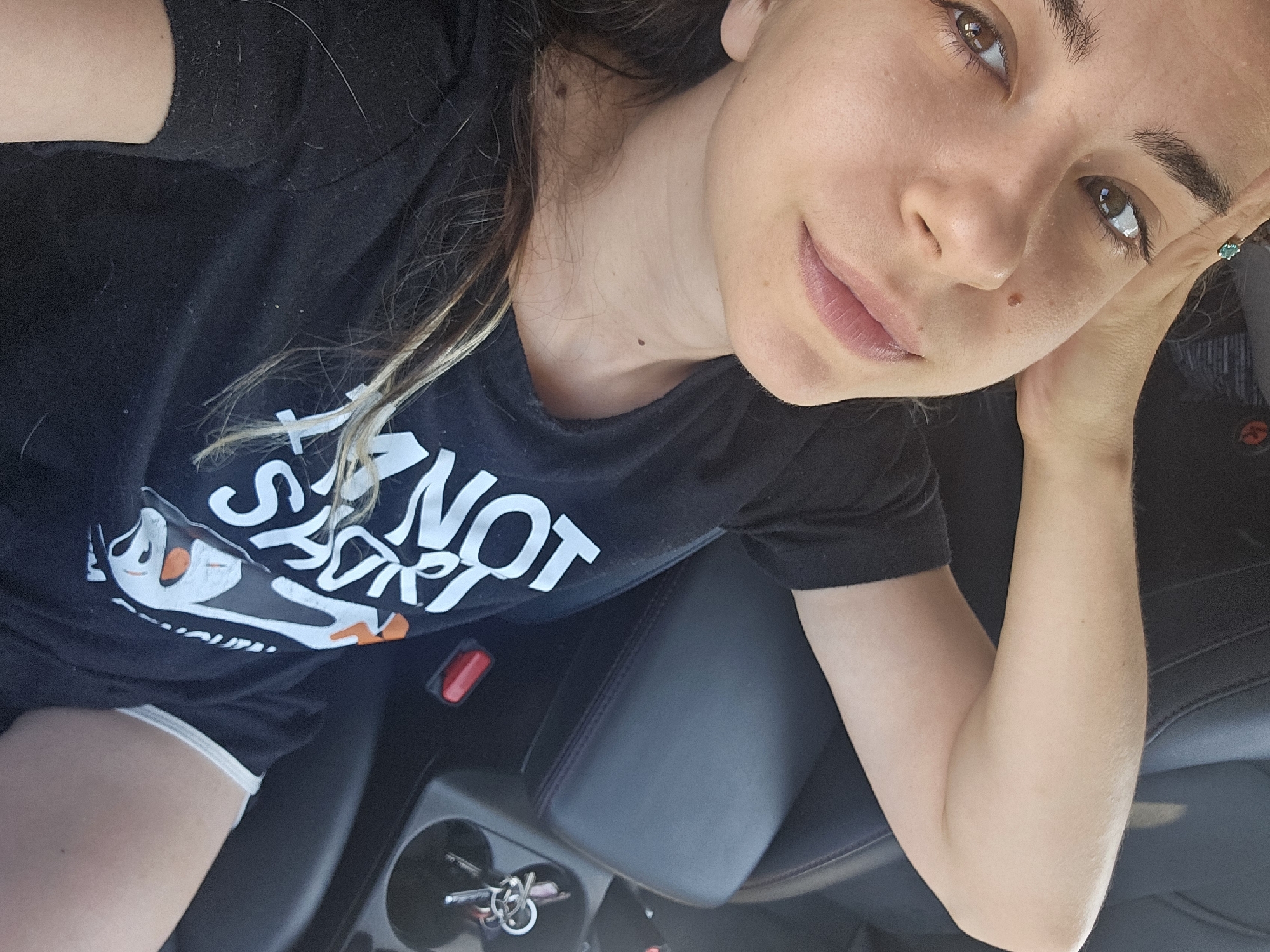 This is a selfie taken by a young woman sitting in the driver's seat of a car. She is wearing a black t-shirt with white text and graphics, and dark shorts with white trim. Her right arm is resting on the center console of the car, and her left hand is holding her face near her cheek. Her eyes are looking up and to the left, and she has a slight smile on her face. The interior of the car is visible, including the center console with a cup holder and keys, and the car seat. The lighting suggests it is daytime, possibly late afternoon, given the warm, natural light illuminating her face. The woman appears relaxed and comfortable. The text on her shirt reads "I'M NOT SAVING." The location is identified as Timi Community, Cyprus, though no explicit visual cues in the image confirm this.