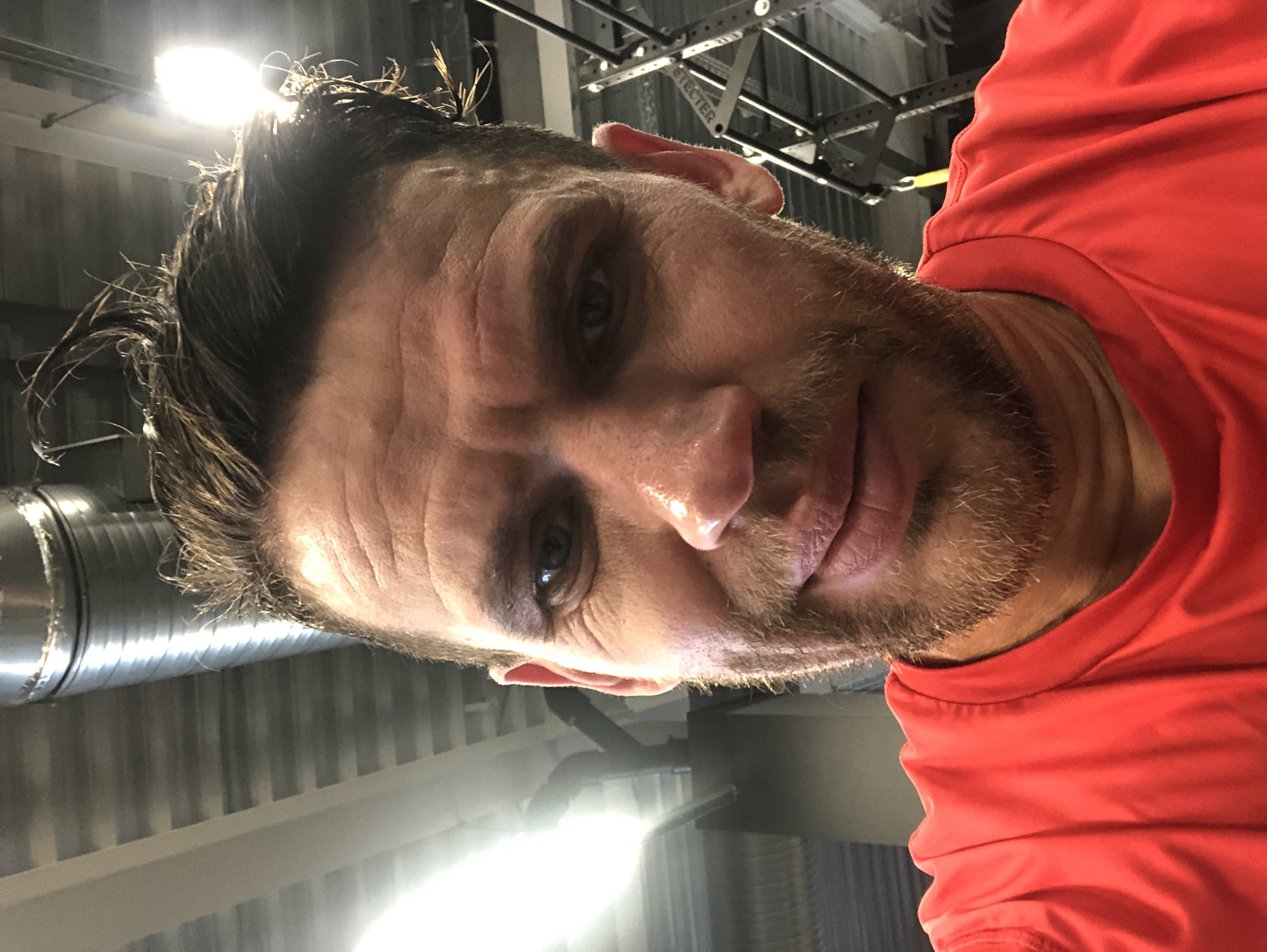 The image is a close-up, selfie-style photograph of a man's face and upper chest, taken indoors. The man has light brown hair, which appears slightly damp and styled upwards. He has visible wrinkles on his forehead and around his eyes, suggesting his age may be between 40 and 50. He has stubble on his chin and upper lip, forming a short beard. His skin appears flushed, and there is a sheen of sweat on his forehead and nose, indicating he may have been exercising. He is wearing a bright red t-shirt. The background is out of focus but suggests an industrial or gym environment with metal structures and lighting. There are no identifying landmarks visible that would indicate a specific location in Saint Petersburg, Russia.