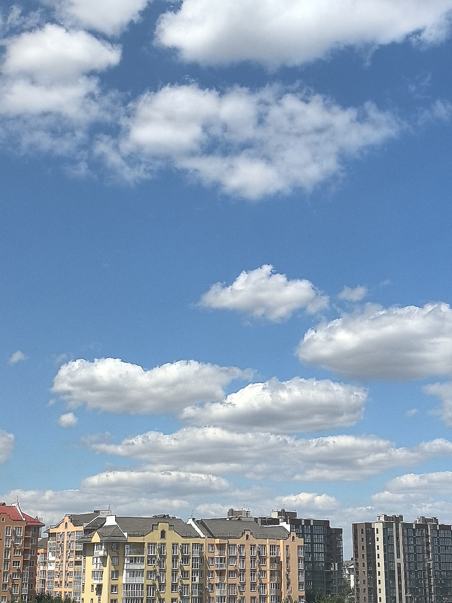 The image depicts a bright, sunny day with a clear blue sky dotted with fluffy white clouds. In the foreground, a row of apartment buildings with varied architectural styles and colors stretches across the frame. The buildings are multi-story structures with numerous windows. To the left, a building with a red roof is visible. The middle section features buildings in shades of orange and yellow, with some modern, darker-colored buildings with many windows on the right. Trees are partially visible at the bottom edge of the frame, suggesting an urban or suburban setting. The scene appears to be a residential area. The image quality is moderate, with some digital noise.