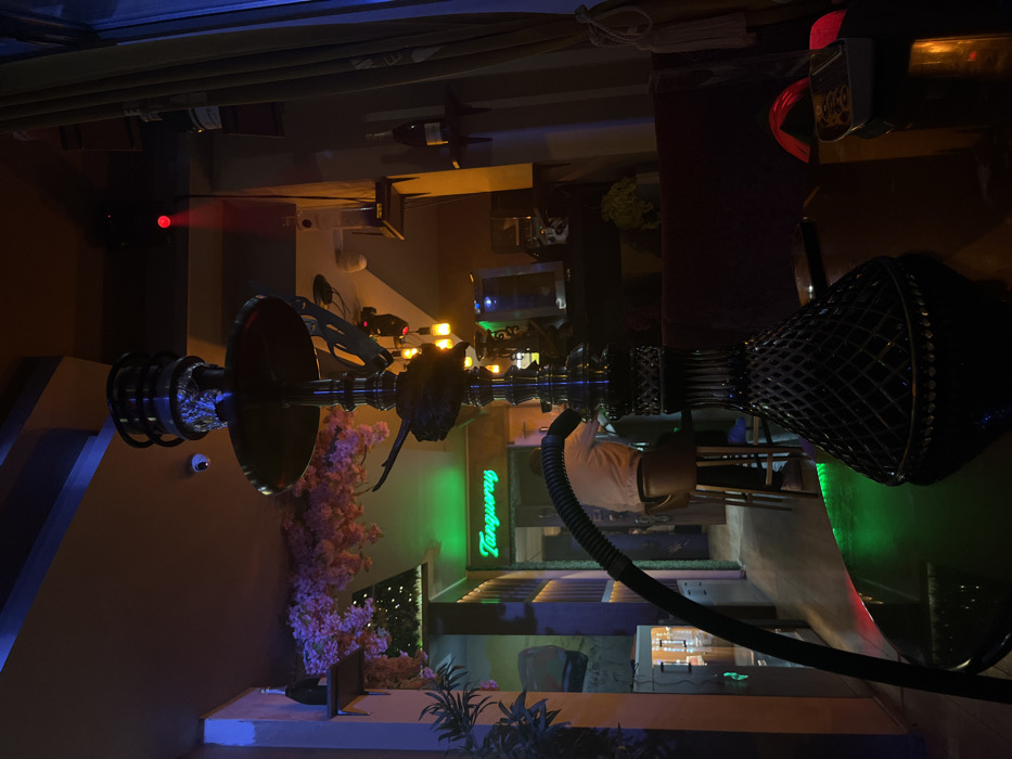 The image displays the interior of a dimly lit shisha lounge or bar in Sofia, Bulgaria, identified by a prominent green neon sign displaying "Tanqueray" in the mid-ground.
In the foreground, a dark-colored hookah stands centrally on a reflective black table. The hookah features a faceted glass base, a metallic stem, and a bowl wrapped in aluminum foil at its apex. A black corrugated hose extends from the stem towards the left. To the right of the hookah's base, a white rectangular object with an imprinted pattern rests on the table. The table surface reflects ambient colored lights, showing areas of green and red.
In the mid-ground, partially obscured, a person is seated on a tall, dark brown stool, facing away from the camera. The individual is wearing a light-colored long-sleeved shirt. Behind this person, an illuminated display or cooler is visible, flanked by vertical dark paneling on the wall.
The background reveals various decorative and functional elements. On the left, artificial pink cherry blossoms are suspended from the ceiling above a green plant. A shelf holding a bottle, consistent with champagne, is mounted on the wall below the blossoms. A white dome-shaped security camera is affixed to the ceiling. On the right side, another shelf displays multiple bottles. A section of deep red upholstered seating is partially visible. A beige or light brown textured curtain hangs on the far right, adjacent to a window. Overhead, a red light source projects a distinct red spot onto the ceiling or upper wall. The overall lighting scheme comprises warm ambient illumination interspersed with blue and green accent lights.