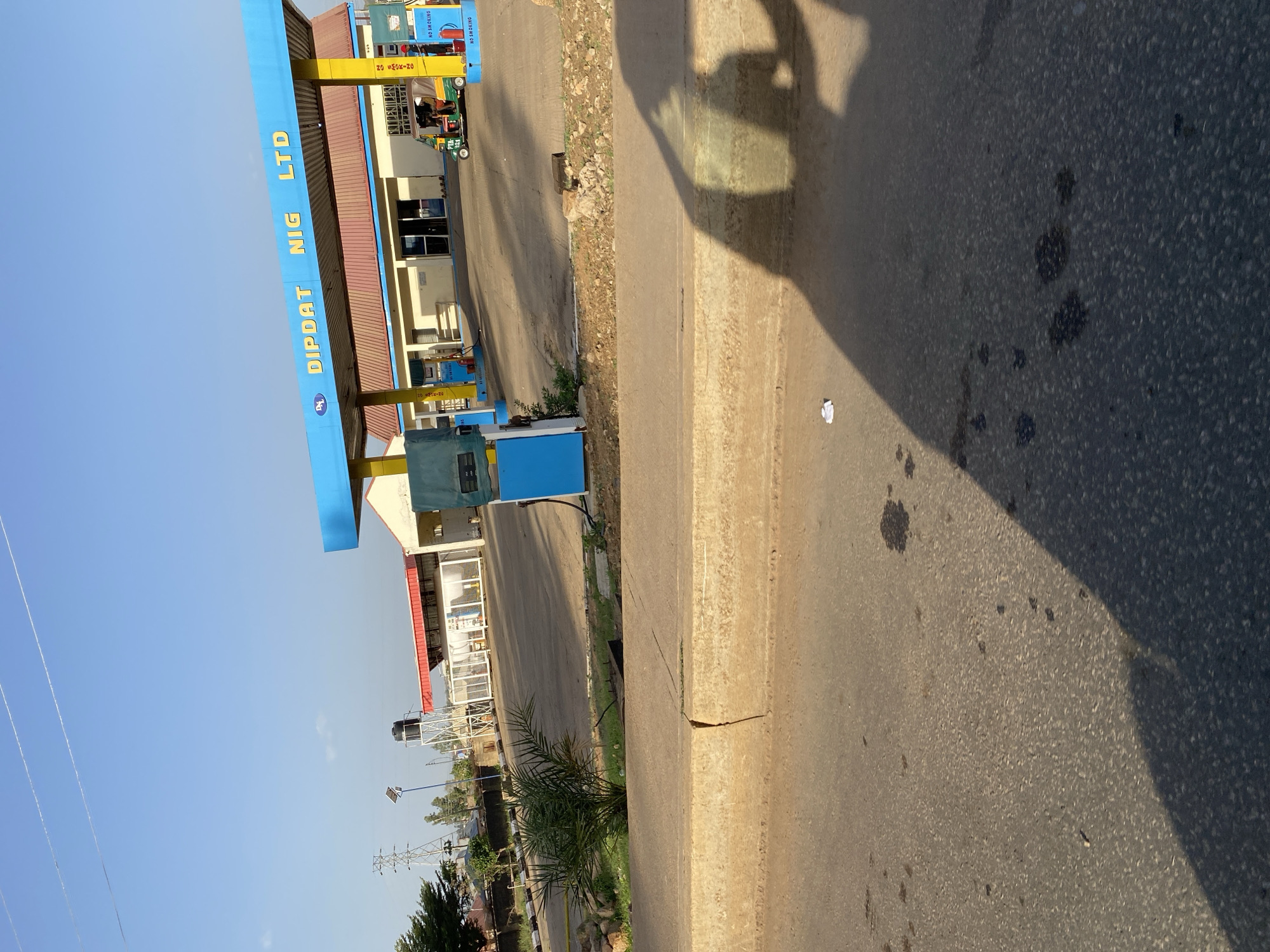 The image shows a Dipdat Nig LTD petrol station in Bukuru, Nigeria. The petrol station has a blue and yellow canopy with the Dipdat Nig LTD name in large yellow letters. There are gas pumps under the canopy, and a small building is attached to the canopy. A yellow three-wheeled vehicle is parked in front of the petrol station. The road in front of the station is paved and has some dark stains. The shadow of a person is visible on the road. In the background are trees, a water tower, and power lines. The sky is clear and blue.