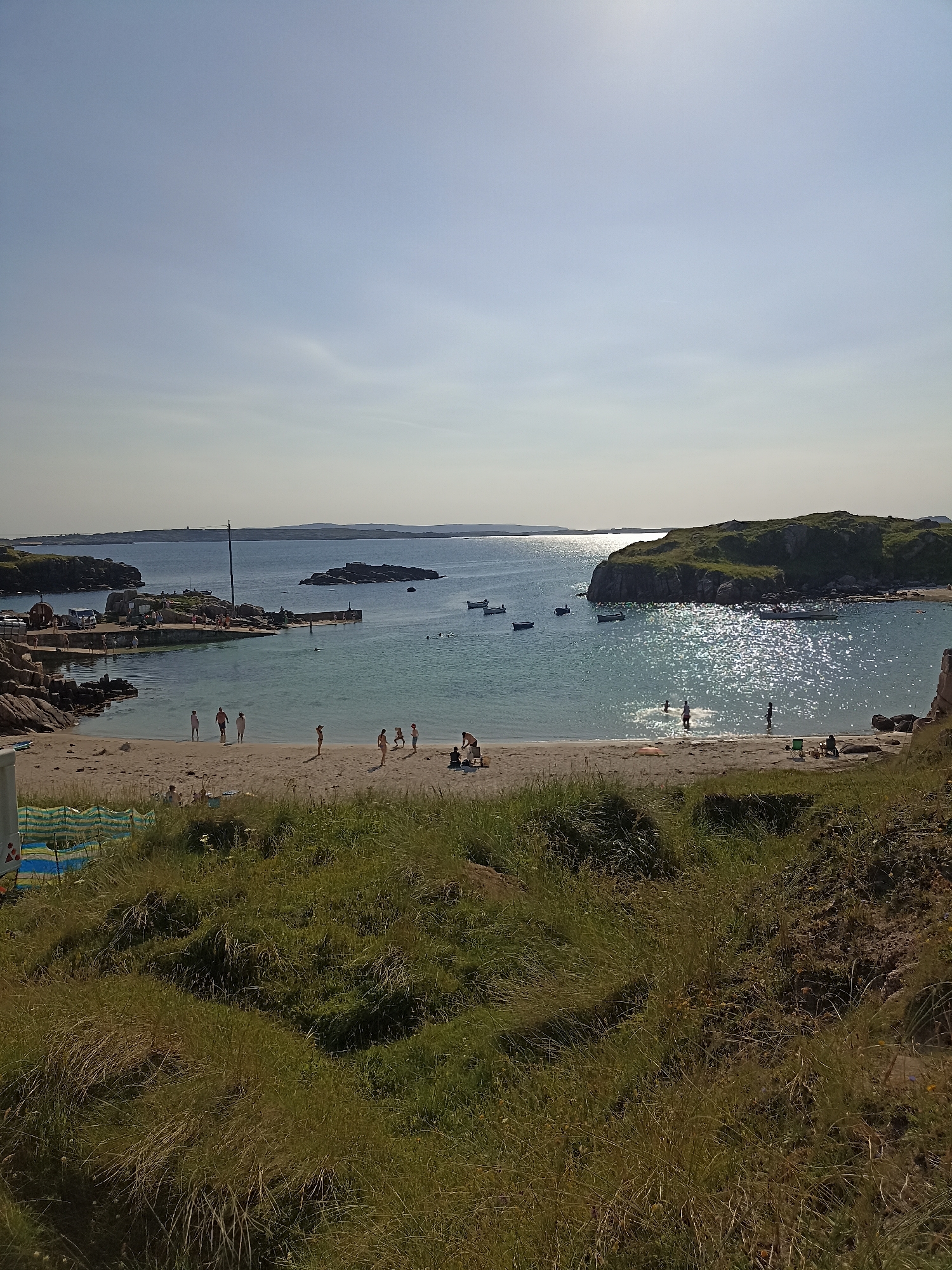 The image depicts a sunny day at a beach in Carnboy, Ireland. The scene is a wide shot of a sandy beach meeting the ocean, with a rocky outcropping on the left and a larger, grassy island in the distance on the right. In the foreground, tall, dry grass covers a slight incline.
On the beach, several people are visible. Closer to the water, a group of children and adults are either standing or walking. Further back on the sand, a few individuals are seated or interacting with each other. The water is clear and calm, with several small boats anchored in the bay. Some of these boats are near the shore, while others are further out towards the distant islands.
To the left of the image, there is a stone pier with a few people on it. Adjacent to the pier, there's a small building and some parked vehicles, suggesting a small harbor or port area. The sky is a clear, pale blue with a slight haziness near the horizon. The sun is bright, casting a shimmering reflection on the water's surface. The overall atmosphere is one of a relaxed summer day at the coast.