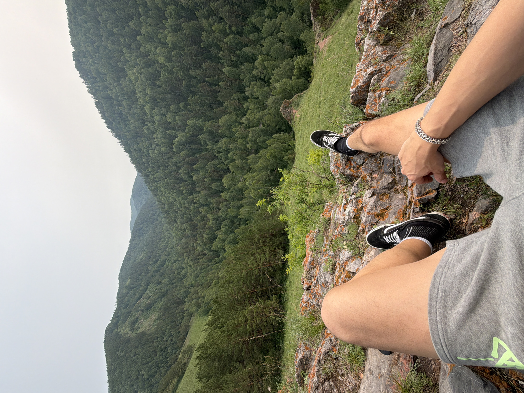 The image depicts a first-person perspective of a person sitting on a rocky outcrop overlooking a heavily forested mountain valley. The person's legs, clad in grey shorts and black sneakers with white laces, are prominent in the foreground. One leg is bent, with the foot resting on a rock, while the other is extended. A silver bracelet adorns the person's wrist. The surrounding environment is characterized by rugged, orange-tinged rocks, green grass, and sparse vegetation. Beyond the immediate foreground, the landscape unfolds into a vast expanse of dense, green forest covering rolling hills and mountains. The sky is overcast and hazy, suggesting a temperate climate. The scene is consistent with the mountainous terrain found in the vicinity of Krasnoyarsk, Russia, known for its expansive forests and natural landscapes.