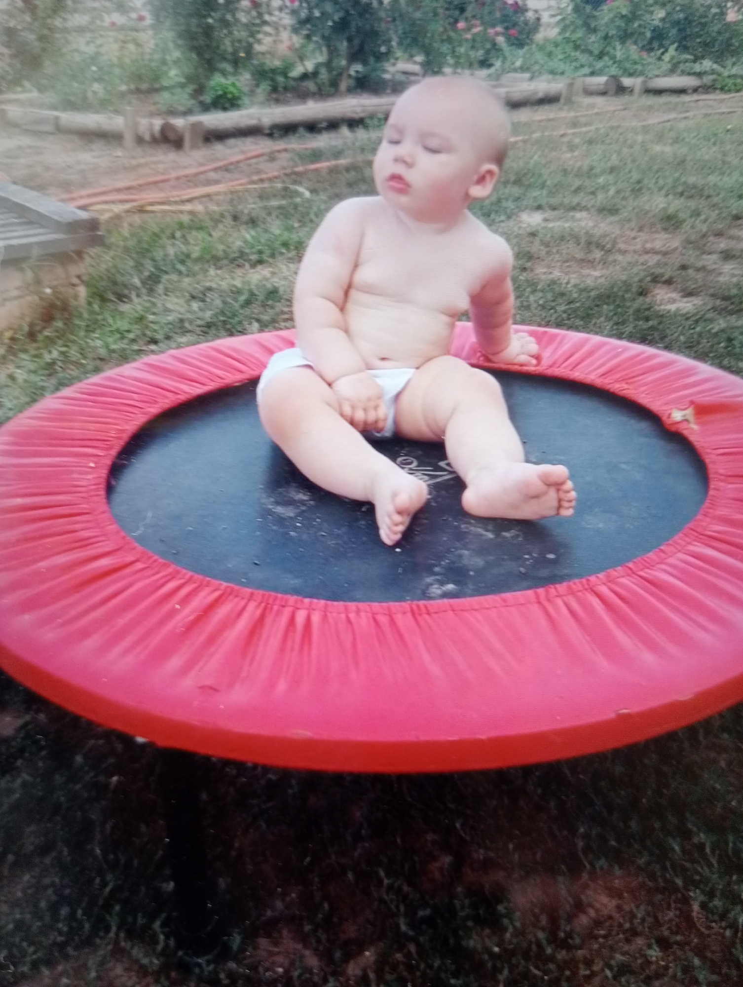 A toddler, with fair skin and light hair, sits on a black trampoline with a red padded edge. The toddler is wearing a white diaper. The trampoline is outdoors, surrounded by grass and dirt. In the background, there are some plants and a wooden log. The image is taken from a slightly elevated angle.