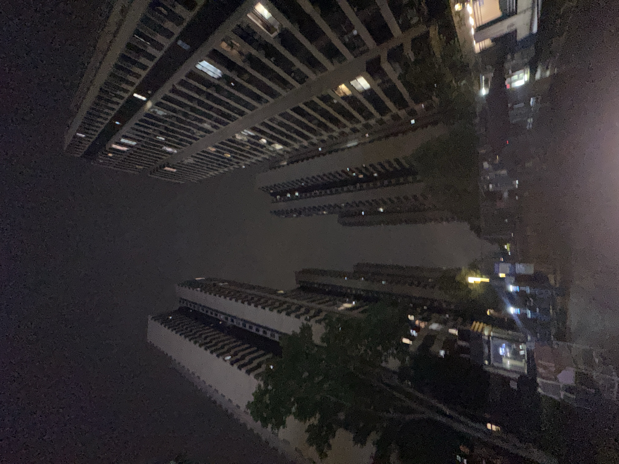 The image depicts a nighttime view of buildings in what appears to be the Chongqing High-tech Zone, China. The perspective is angled upward, showing a cluster of high-rise buildings with lit windows. Tree branches partially obscure the view of some buildings. The sky is dark, suggesting a night scene. The overall lighting is low, with artificial lights from the buildings providing the main illumination.