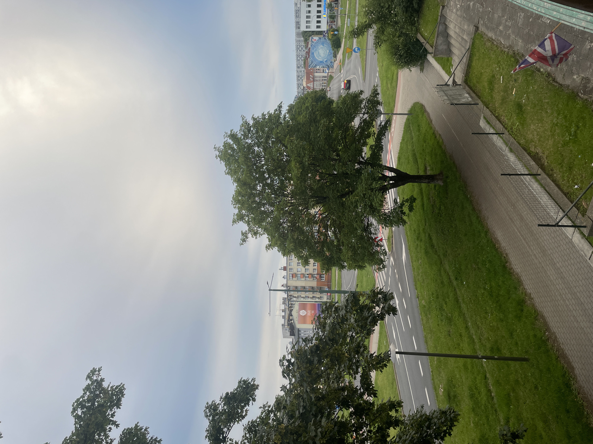 The image captures an overcast day in a residential area of Kołobrzeg, Poland. In the foreground, a paved pedestrian walkway runs alongside a grassy verge, bordered by a metal fence. A British flag is visible on the right side, attached to a railing. Beyond the walkway, a tree-lined street is present, with a road featuring white lane markings and a pedestrian crossing. In the background, several multi-story buildings are visible, some adorned with colorful murals. A single red car is navigating the intersection. The sky is predominantly gray with some diffused sunlight.