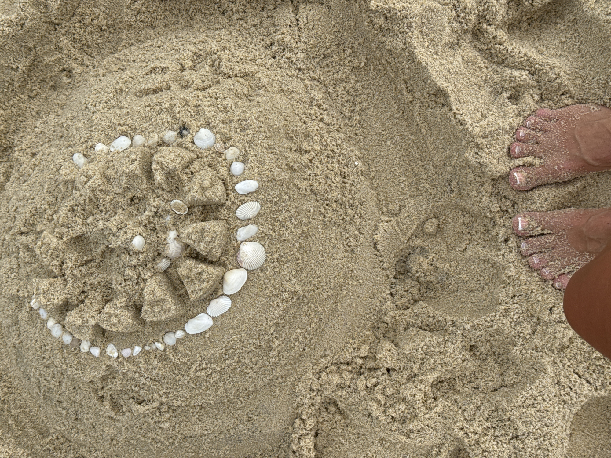 A close-up, high-angle shot shows a circular pattern of small seashells embedded in wet sand. The pattern is incomplete, with gaps and some larger sand mounds within the circle. The sand is textured with fine grains and appears slightly damp, suggesting a beach environment. In the foreground, the tops of two bare feet are visible, partially covered in sand, with painted toenails. The feet are positioned with toes pointing upwards. This scene is located on a beach on Tengah Island, Malaysia.