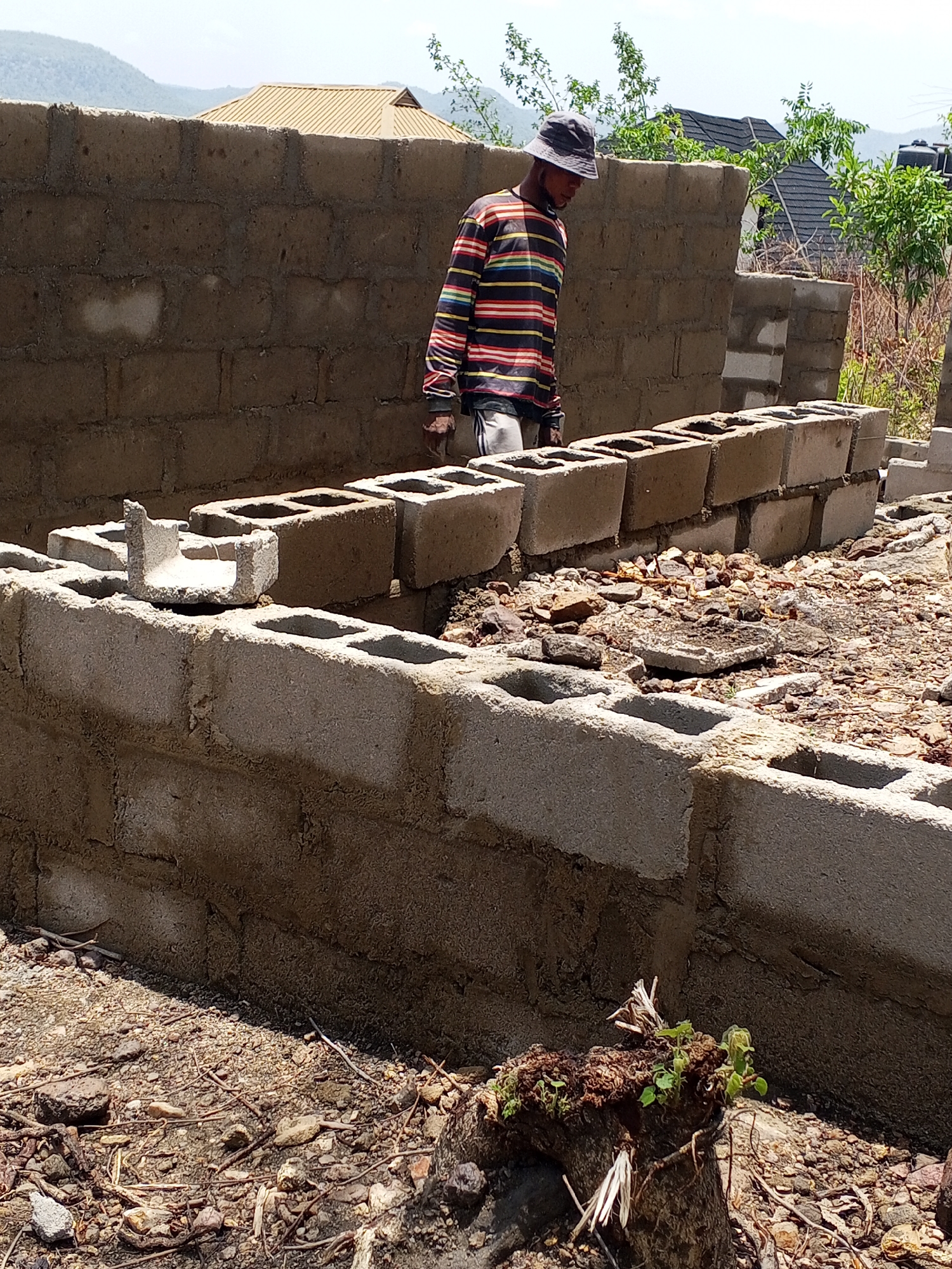 The image depicts a construction site in Lokoja, Nigeria. A partially constructed concrete block wall forms the foreground, with a man wearing a striped long-sleeved shirt and a bucket hat standing behind it. The man is likely involved in the construction process. The foundation consists of a layer of concrete blocks. Debris and rocks are scattered on the ground. In the background, there is a partially constructed structure and other houses. The background shows mountainous terrain and vegetation. The time of day appears to be daytime, based on the light.