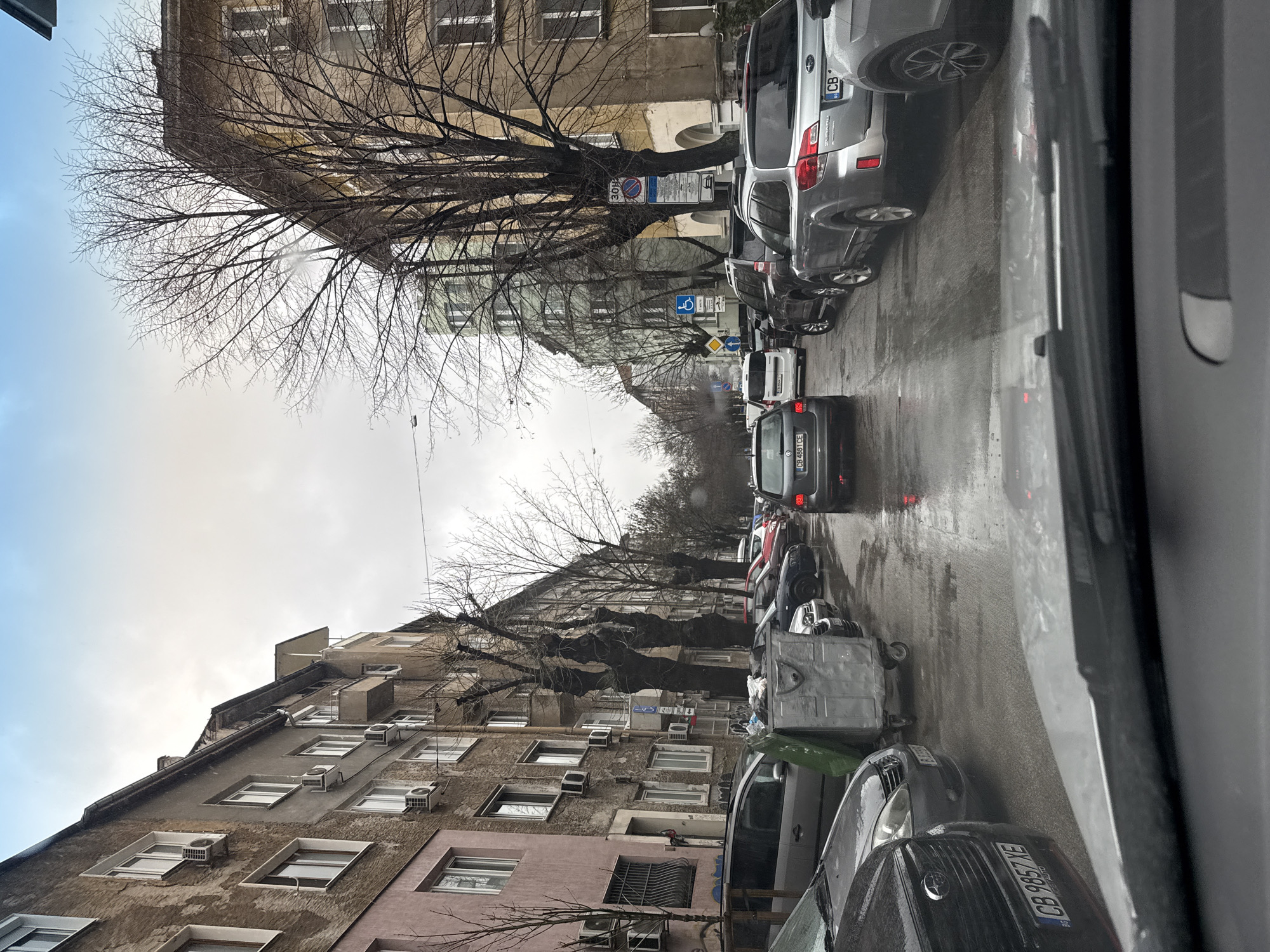 The image presents a forensic view of an urban street in Sofia, Bulgaria, observed from inside a vehicle through a wet windshield on an overcast day. The wet asphalt street is densely populated with parked and moving vehicles. On the left side, a series of multi-story residential buildings with weathered facades, varying in shades of pink, brown, and grey, feature numerous external air conditioning units and windows. A large grey industrial dumpster with wheels is positioned on the left curb between parked cars. On the right side, similar multi-story buildings, some painted yellow/ochre, are partially obscured by bare deciduous trees with dark, gnarled trunks. Several traffic signs are visible on the right, including a circular blue sign indicating a "30H" speed zone, a blue square sign with a white wheelchair symbol, and a yellow diamond-shaped priority road sign. Numerous cars are present, including a silver Subaru Outback SUV on the right and a dark sedan directly ahead with illuminated brake lights. License plates visible on multiple vehicles prominently display the "CB" prefix, confirming the location as Sofia. No individuals are visible within the frame.