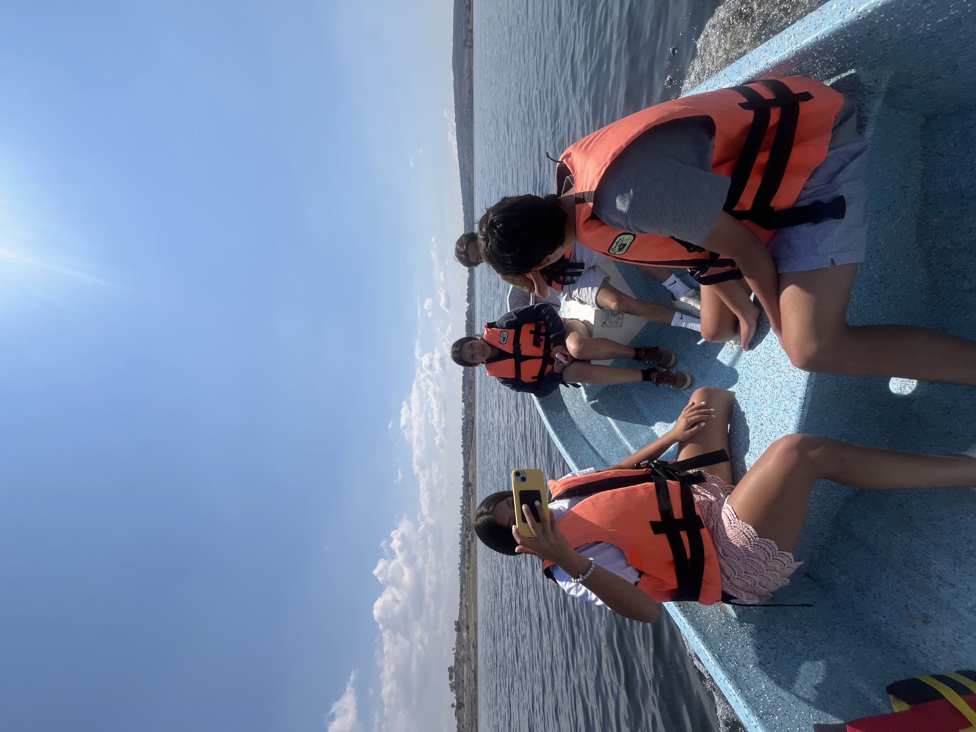 The image shows a group of people on a boat on a lake in the city of El Oasis Valsequillo, Mexico. The people are wearing life jackets and appear to be taking a boat trip. One person is holding a cell phone. The lake water is dark and rippled, and the sky is clear with a few clouds. A strip of land can be seen in the background.