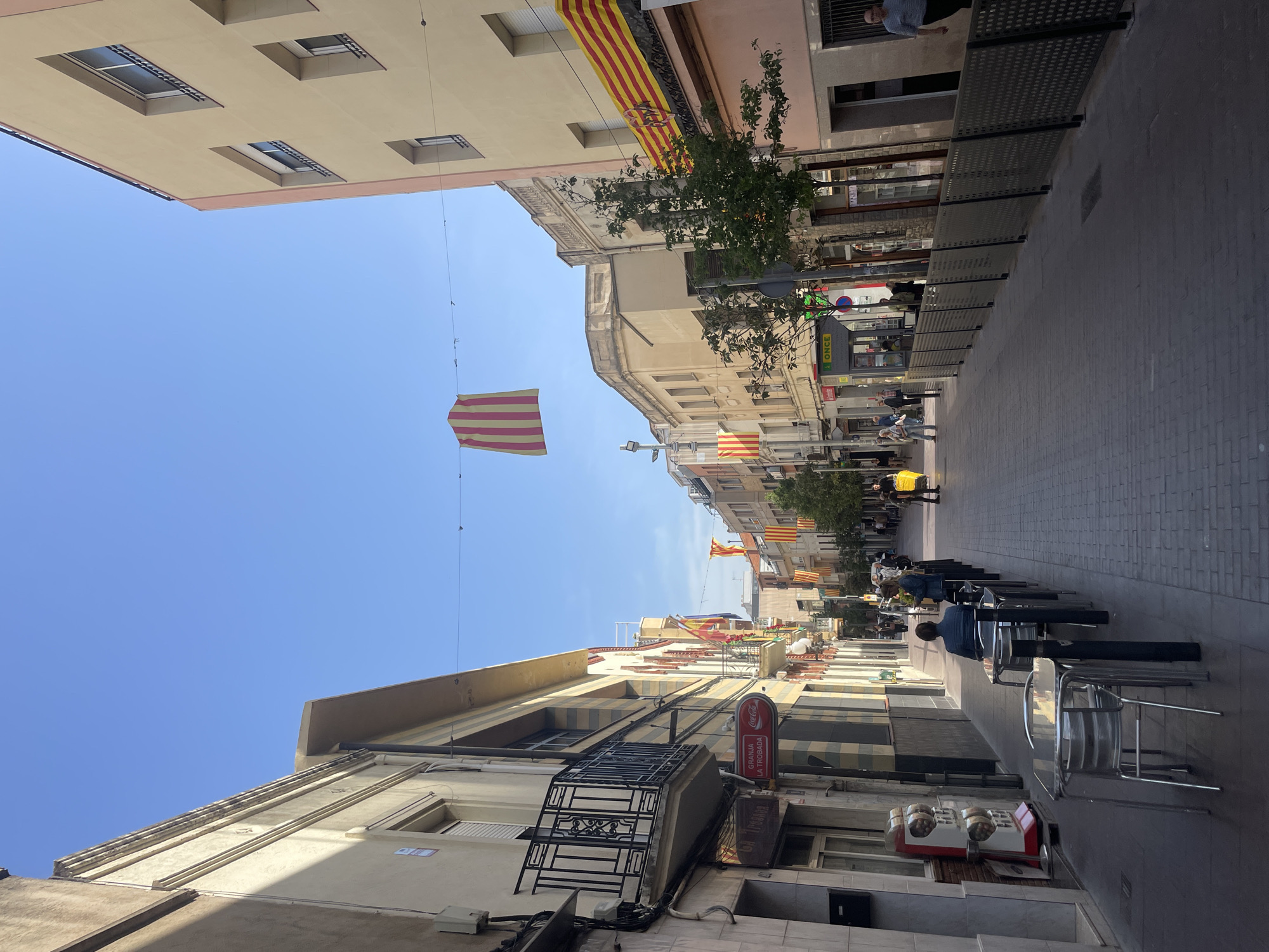 The image depicts a pedestrian street in Montcada, Spain, characterized by buildings on either side decorated with Catalan flags (red and yellow stripes). The sky is clear and blue. Several people are walking along the street, and others are seated at tables outside a cafe or restaurant. A metal grate serves as a barrier or fence along part of the street. The buildings have varied architectural details, including balconies and visible windows. A sign for "Granja La Trobada" can be seen on one of the buildings. The overall scene suggests a typical day in the town.