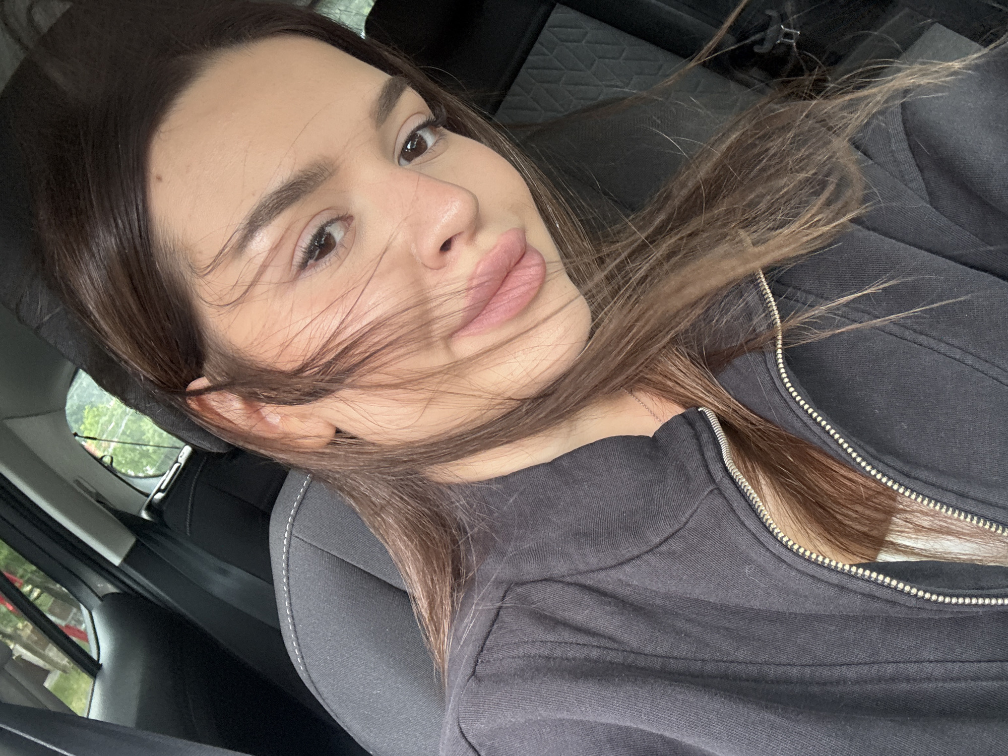 This media file is a close-up, selfie-style photograph of a young woman sitting in the passenger seat of a car. She has long, dark brown hair that is blowing across her face. The woman is wearing a dark gray zippered jacket. Her eyes are brown, and she is looking towards the camera with a slight smile. The background shows the interior of the car, including a headrest and the side window with greenery visible outside. The lighting suggests it is daytime, and the overall mood appears casual and relaxed. The scene is set in Prague, Czechia. There is no visible text in the image.