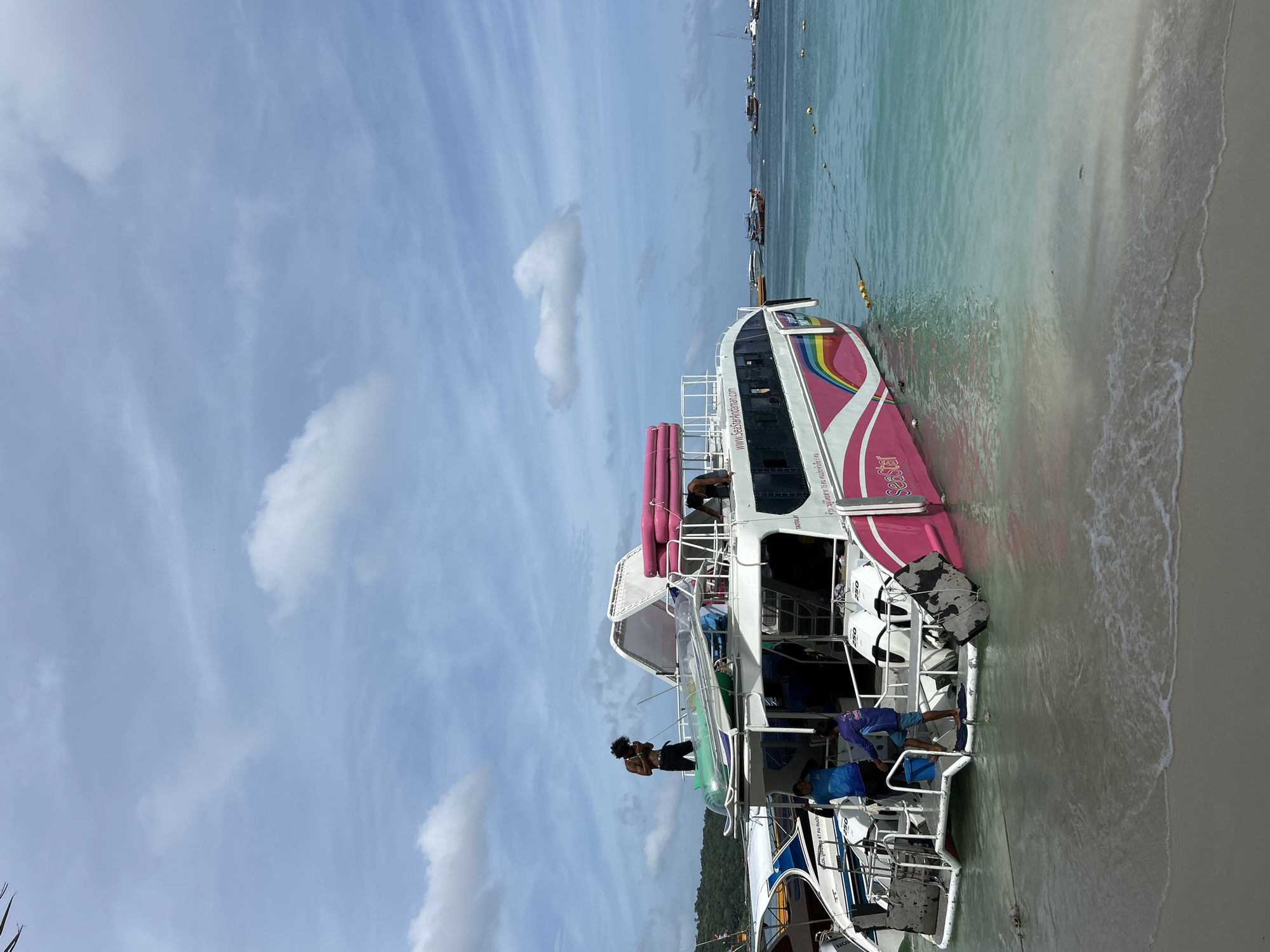 A pink and white catamaran ferry boat is docked on the shore of Ao Nang, Thailand. The boat has a rainbow stripe design on its side and the words "SeaStar" and "www.SeaStarAndaman.com" are visible. Several people are on the boat, some appearing to be crew members and others passengers. One person stands on the top deck of the boat, looking at their phone. Another person is descending the stairs from the upper deck. The water is clear and turquoise, with a sandy beach in the foreground. Other boats are visible in the distance. The sky is blue with scattered white clouds.